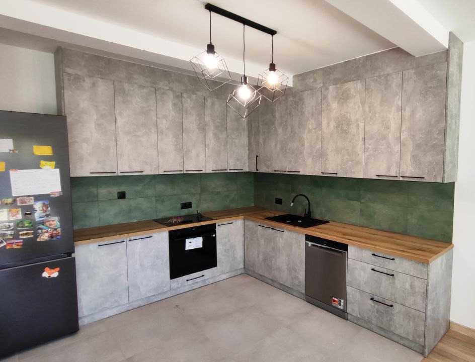Szara kuchnia z blatem imitującym drewno, zielonymi płytkami nad blatem i nowoczesnymi lampami wiszącymi.