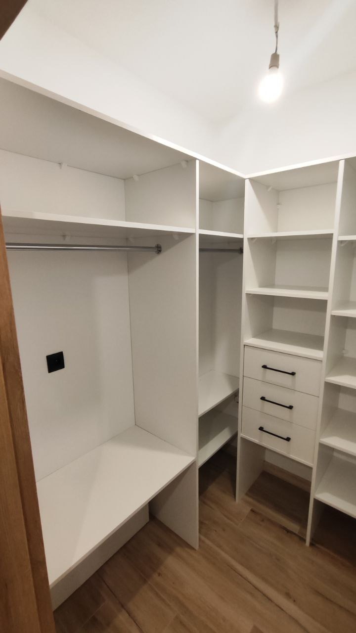 Biała, narożna garderoba z półkami i szufladami z czarnymi uchwytami, oświetlona pojedynczą żarówką, widok z perspektywy wejścia na drewnianą podłogę.