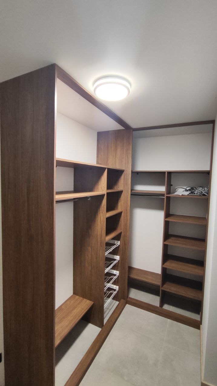 Wnętrze garderoby z drewnianą zabudową, widoczne półki, drążki na ubrania i wysuwane kosze, oświetlenie sufitowe LED.