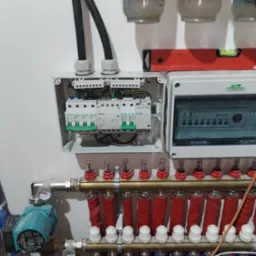 Rozdzielacz ogrzewania podłogowego z pompą obiegową, zabezpieczeniami elektrycznymi w skrzynkach, poziomica na górze, rury w izolacji czerwonej i niebieskiej.