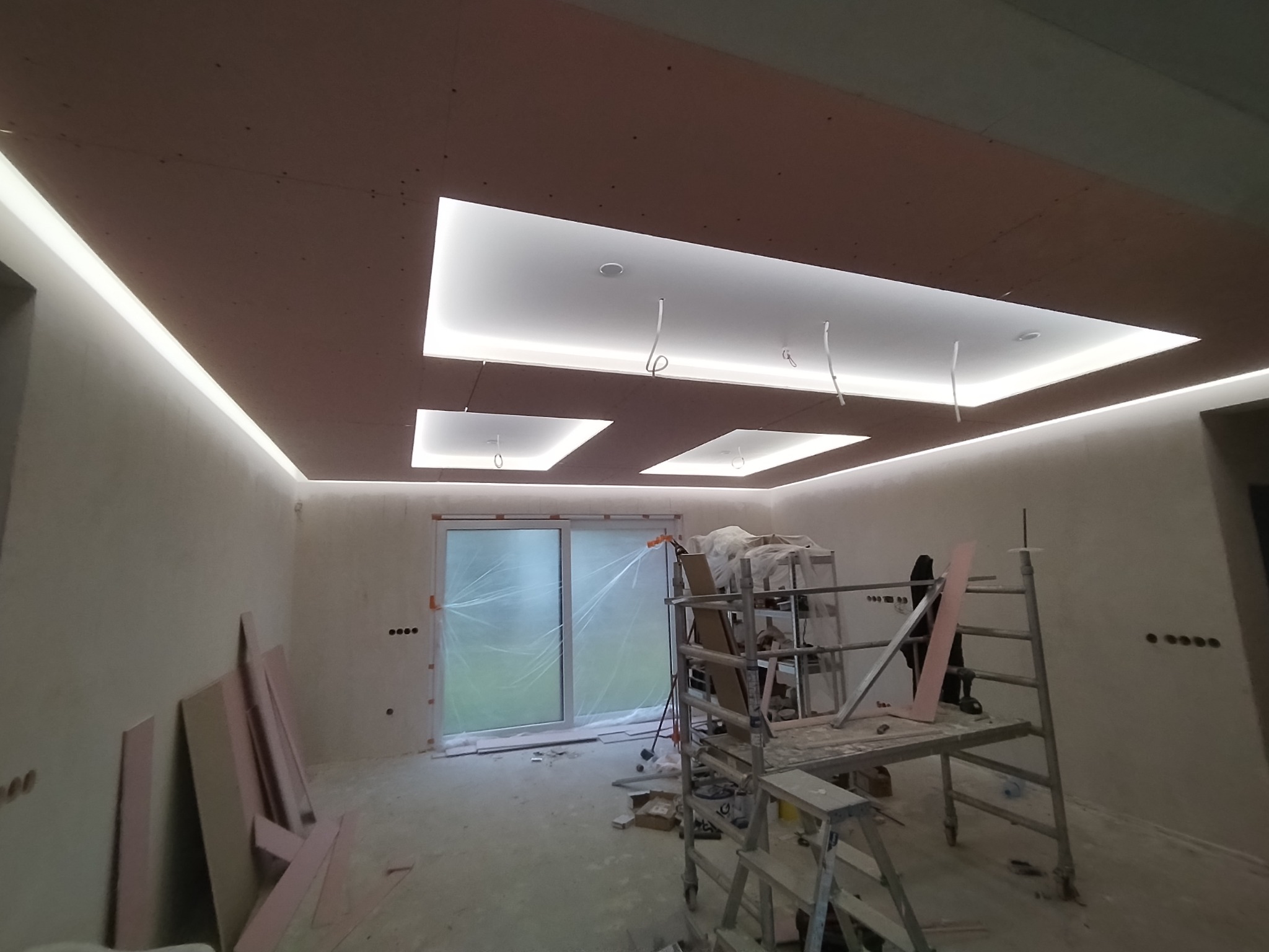 Wnętrze w trakcie wykańczania: sufit z podświetleniem LED i wnękami, rusztowanie, płyty gipsowe oparte o ścianę, folia ochronna na oknach.