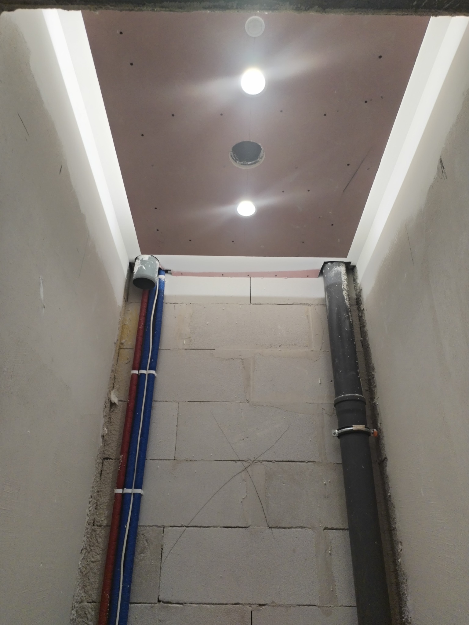 Wnętrze w trakcie remontu: widoczne piony instalacyjne (woda, kanalizacja) przy ścianie z bloczków, podwieszany sufit z trzema otworami na oświetlenie punktowe, instalacja elektryczna w peszlach.