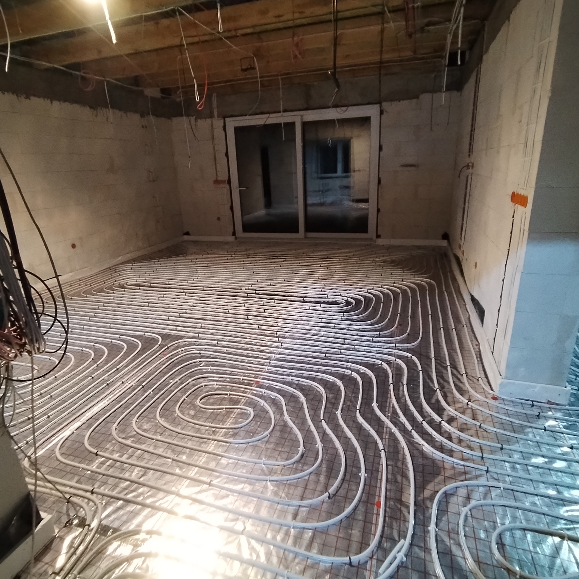 Rozłożone rury ogrzewania podłogowego na siatce zbrojeniowej, widoczne także przewody elektryczne wystające z sufitu w surowym stanie.