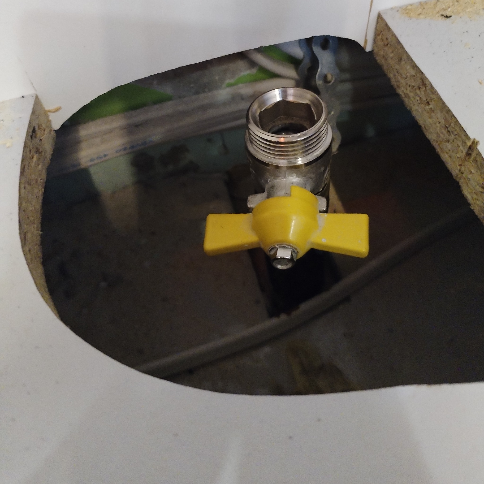 Metalowy zawór gazowy z żółtą rączką wystający z otworu w białej płycie meblowej. Widoczne elementy instalacji w tle.