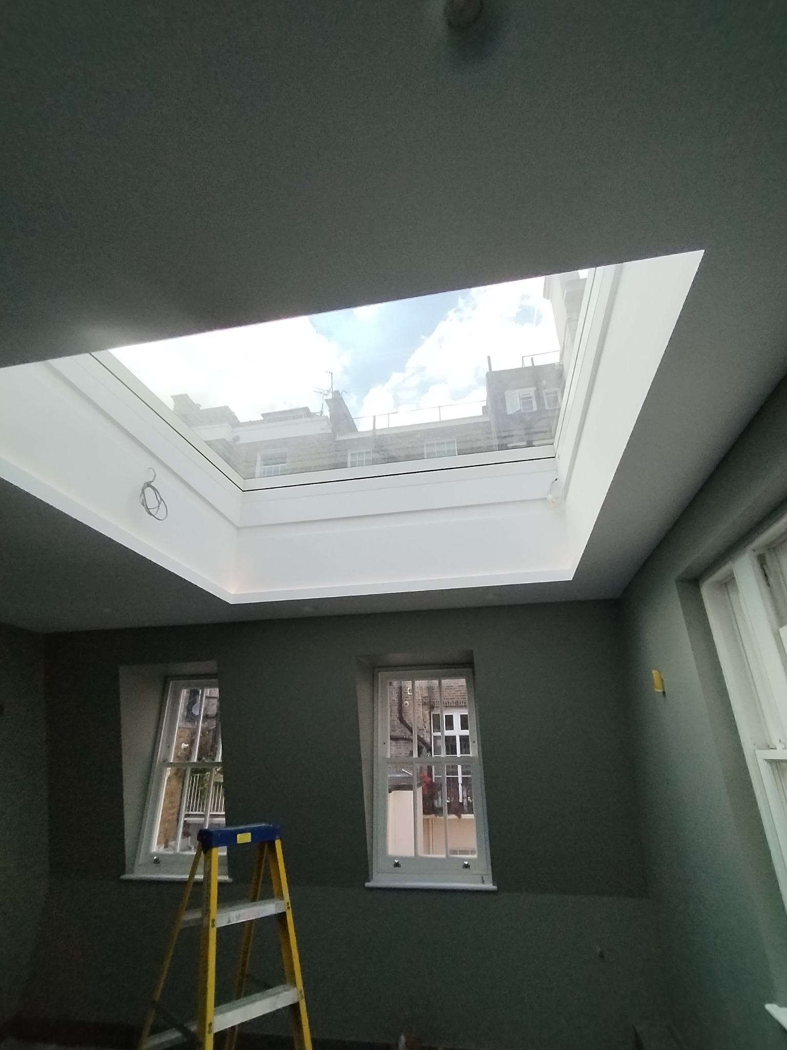 Wnętrze pokoju z widokiem na niebo przez okno dachowe, widoczna drabina oparta o ścianę, dwa okna w ścianie.