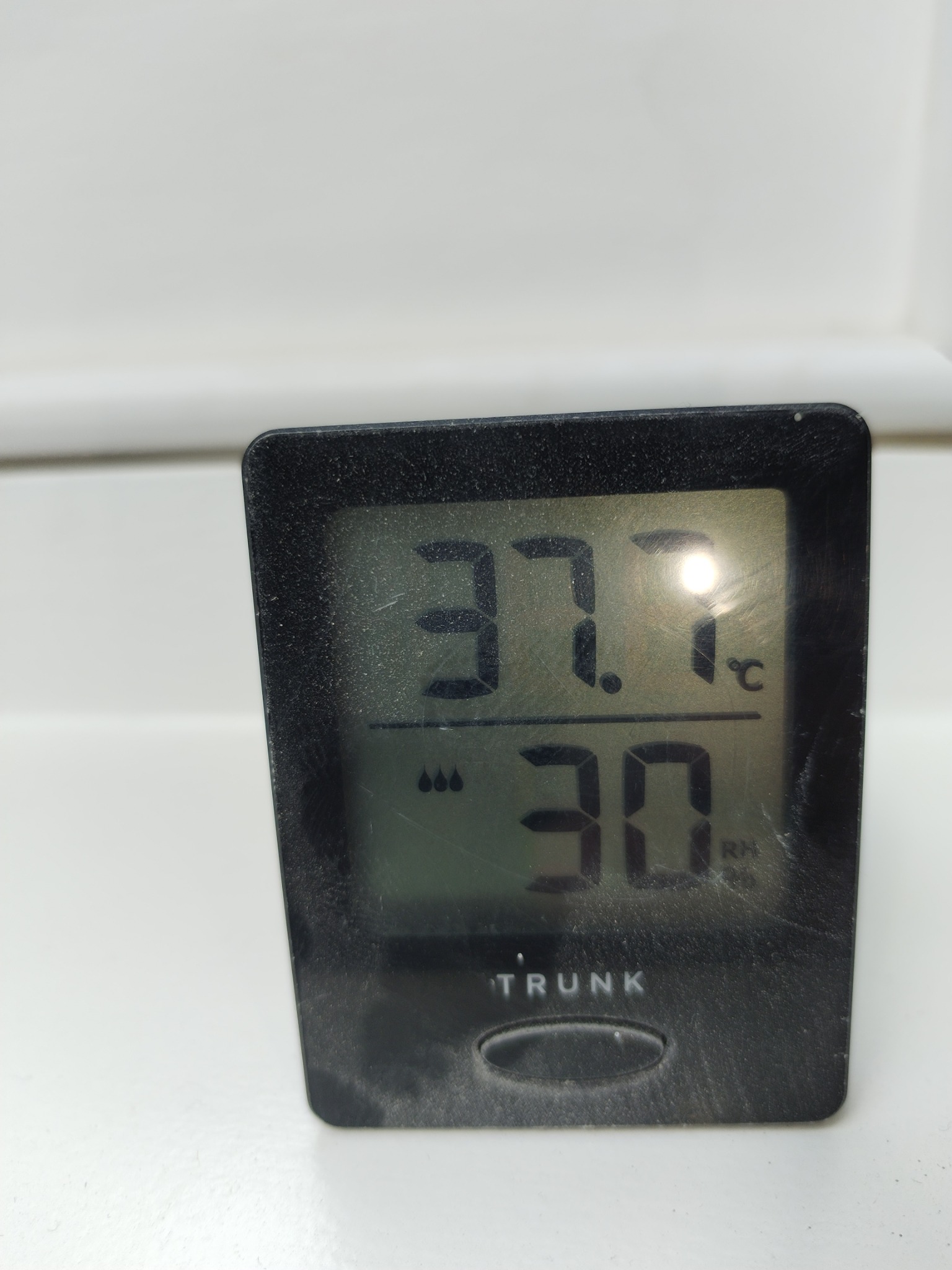 Czarny, zakurzony termometr elektroniczny TRUNK wyświetla temperaturę 37.7 stopni Celsjusza i wilgotność 30%.