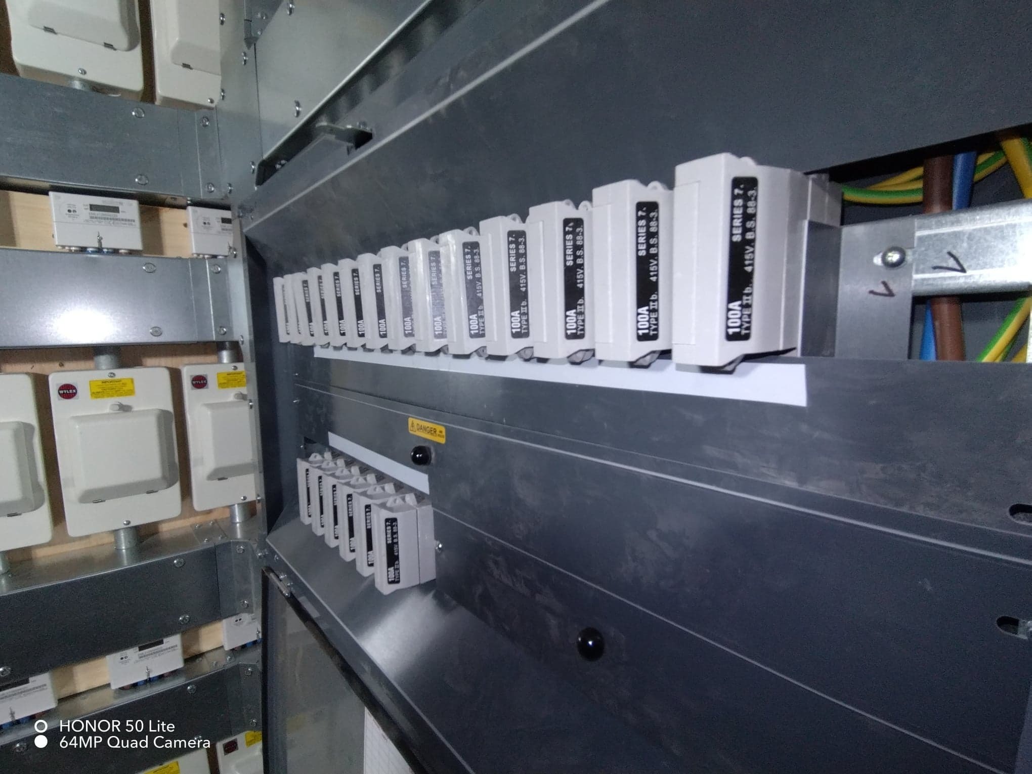 Szyna z zamontowanymi bezpiecznikami 100A w metalowej obudowie rozdzielnicy elektrycznej, widoczne przewody zasilające.