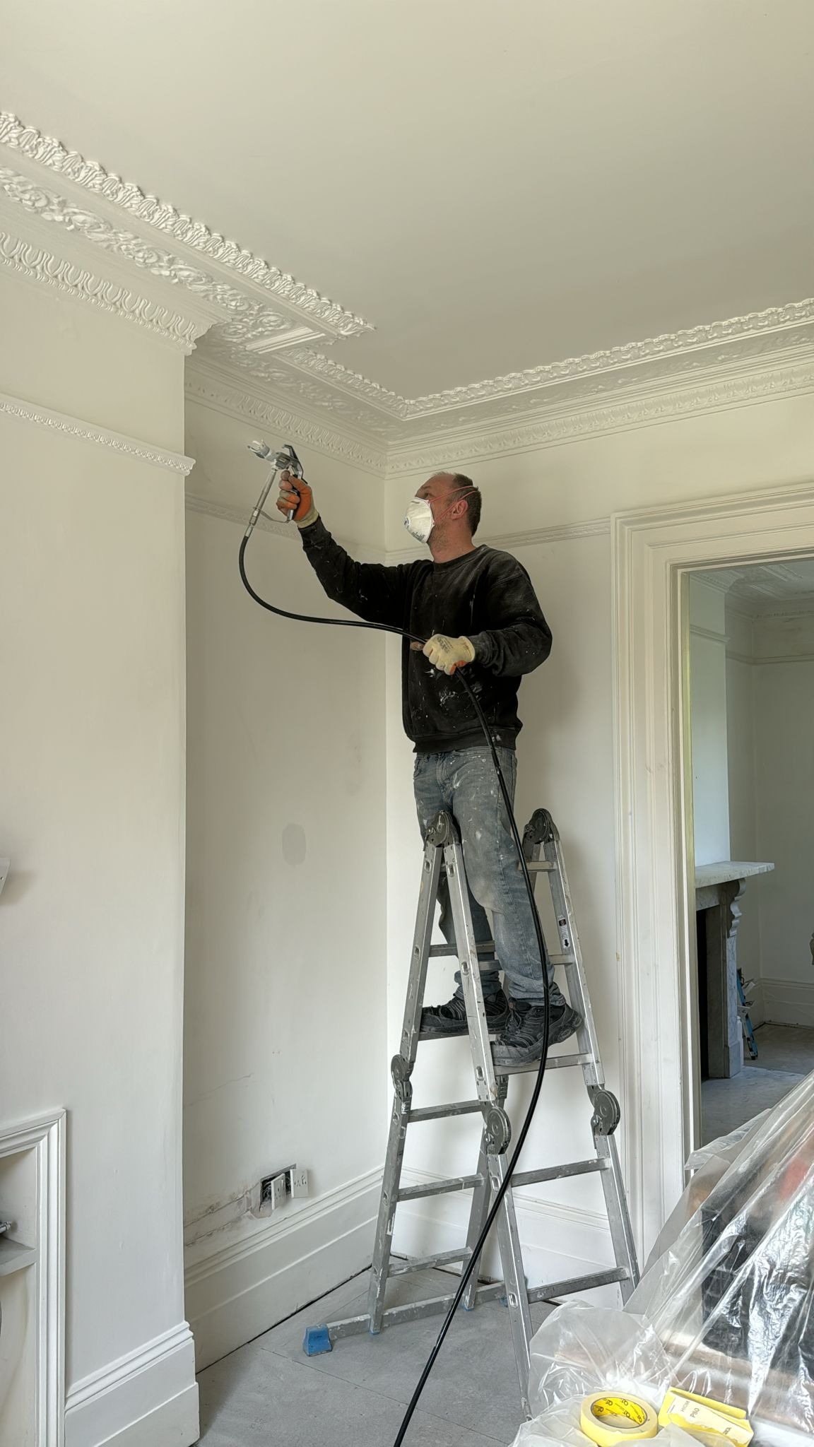 Pracownik w masce maluje natryskowo sufit z ozdobną sztukaterią, stojąc na aluminiowej drabinie w jasnym wnętrzu.