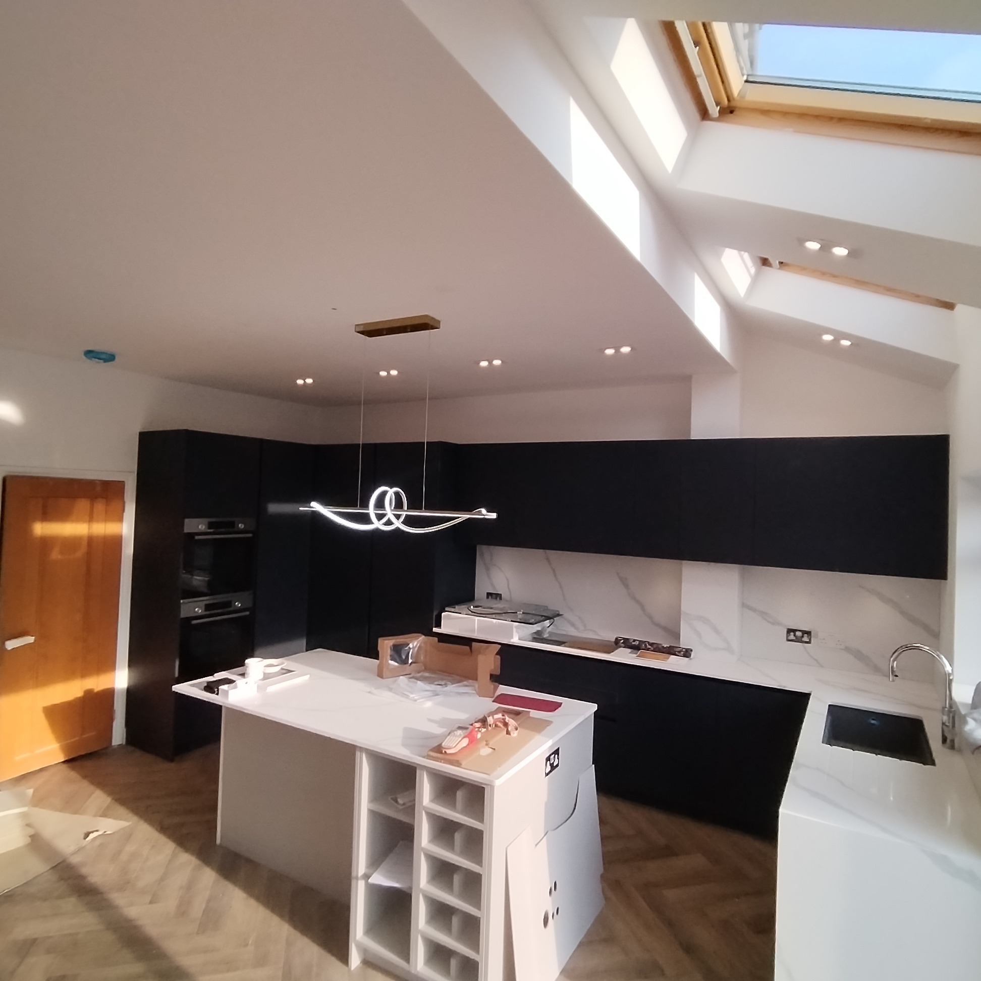 Nowoczesna kuchnia w trakcie montażu: czarne szafki, białe blaty z marmurowym wzorem, wyspa kuchenna z miejscem na wino, designerska lampa wisząca i drewniana podłoga w jodełkę, doświetlona przez...