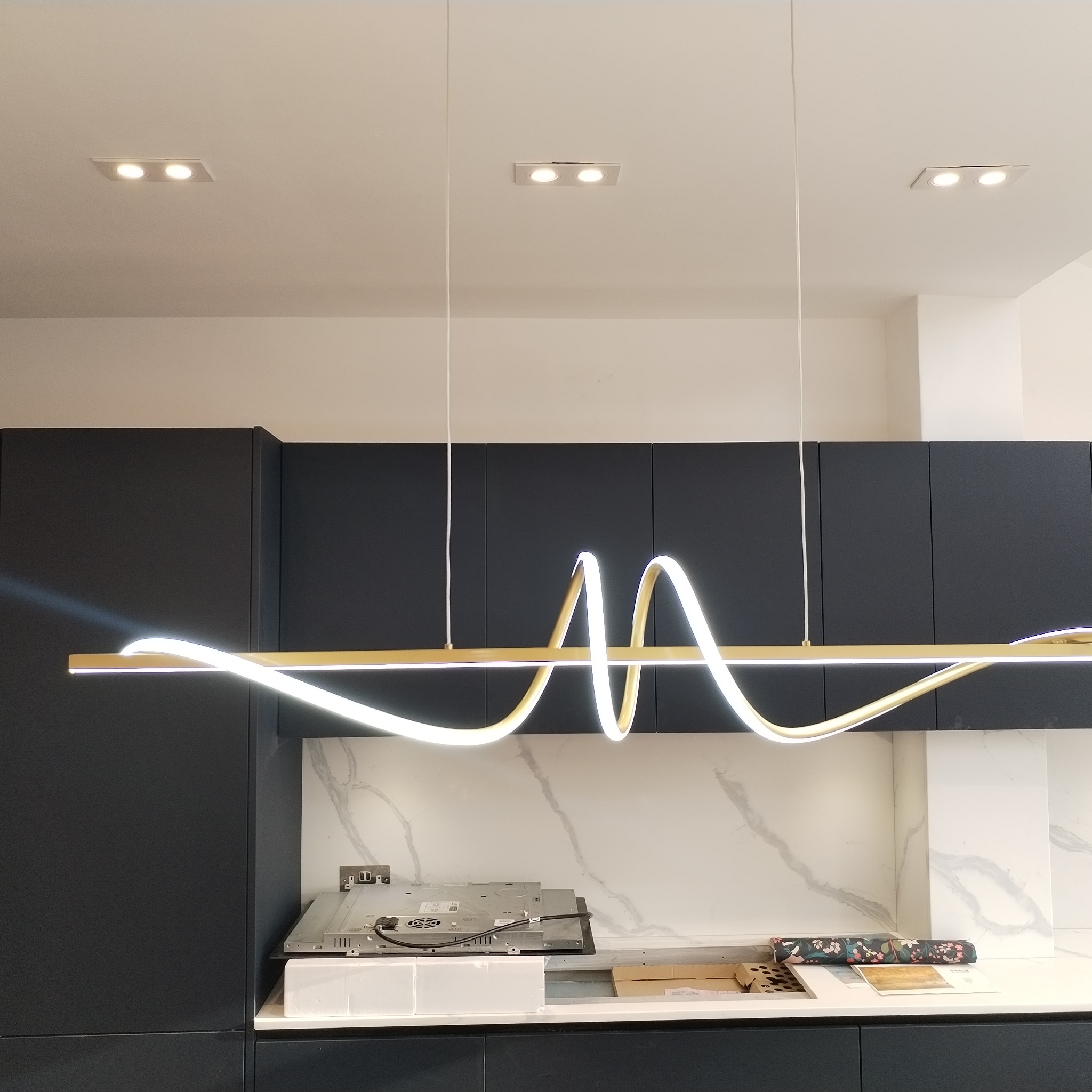 Nowoczesna kuchnia z czarnymi szafkami, marmurowym backsplash'em i designerską lampą LED w kształcie fali nad miejscem przygotowywania posiłków, z widoczną niedokończoną instalacją płyty indukcyjnej.