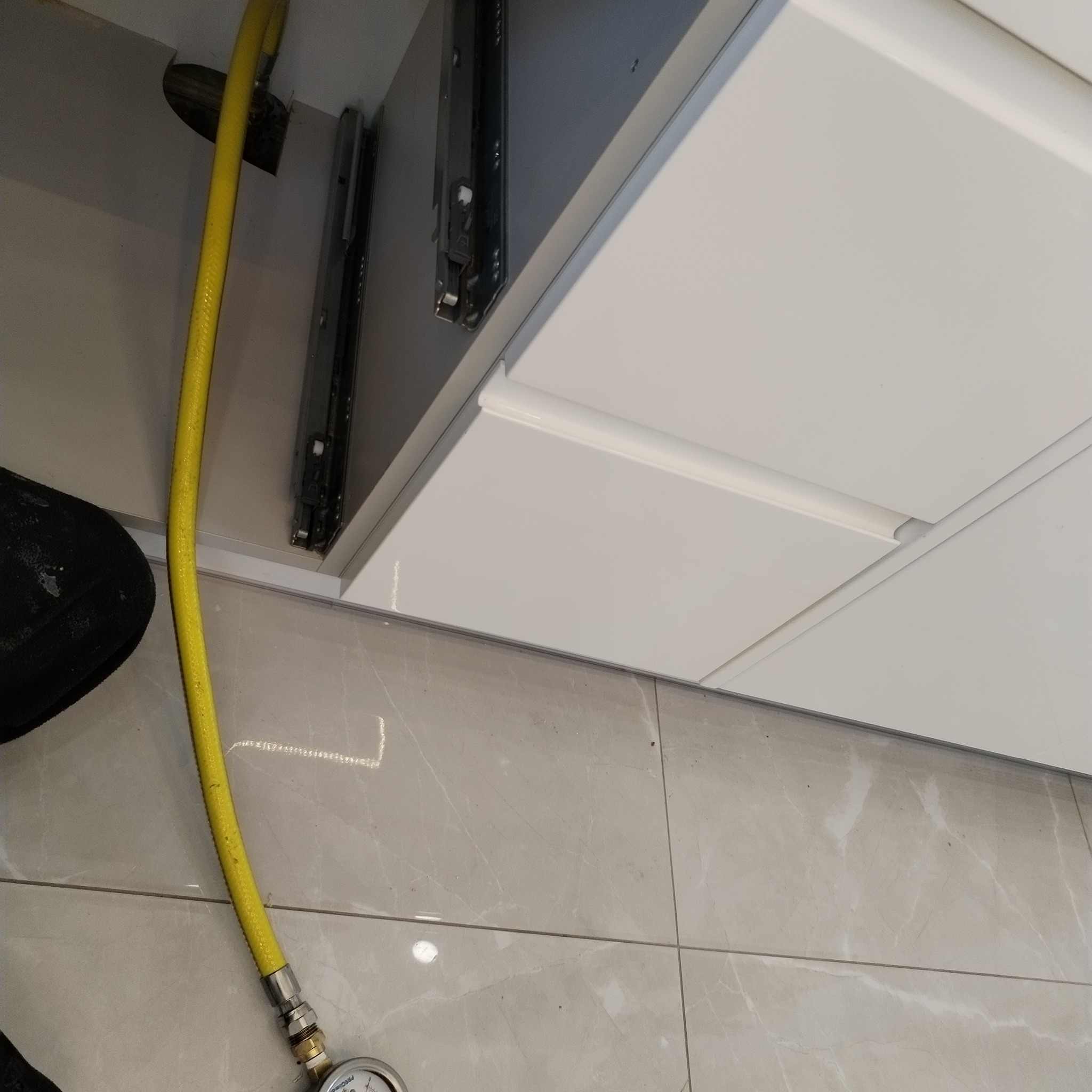 Żółty przewód gazowy podłączony do instalacji w szafce kuchennej, widoczny manometr, szare płytki podłogowe.