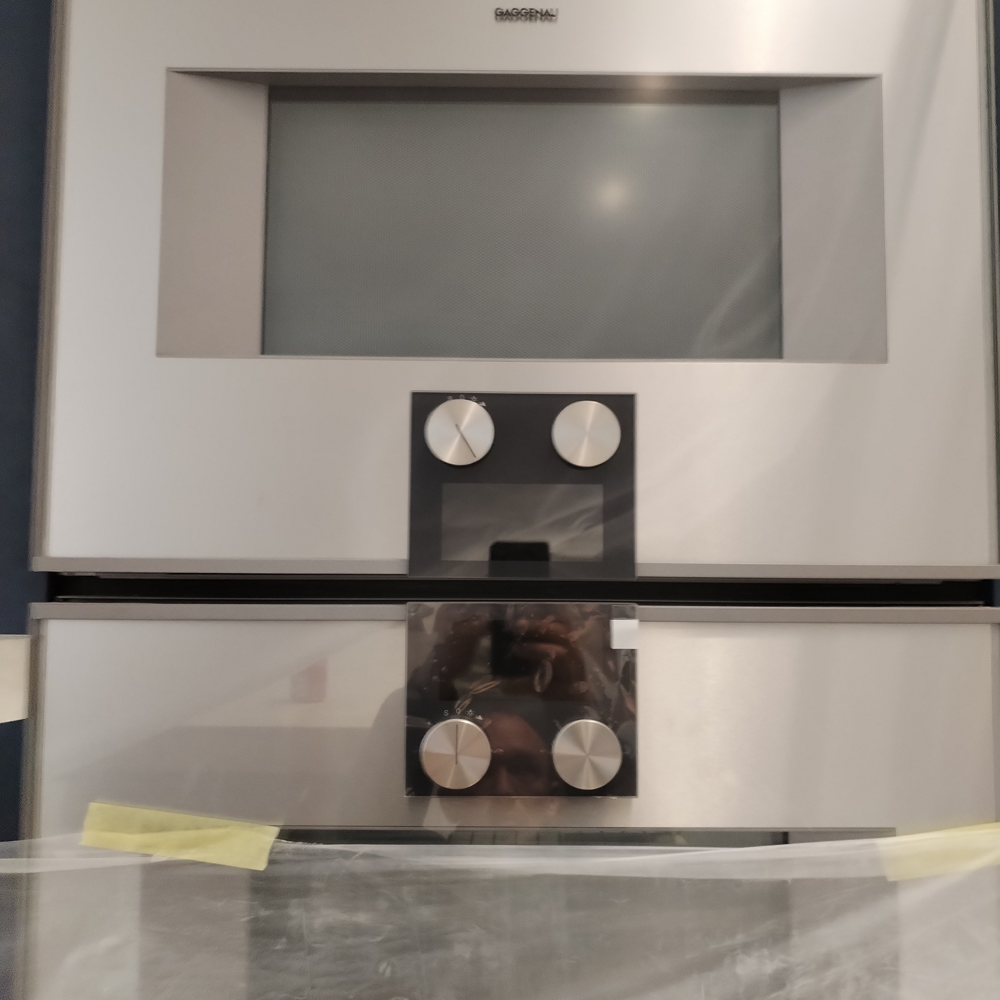 Nowoczesny piekarnik marki Gaggenau, zabezpieczony folią ochronną i taśmą, z widocznymi panelami sterowania i wyświetlaczem.