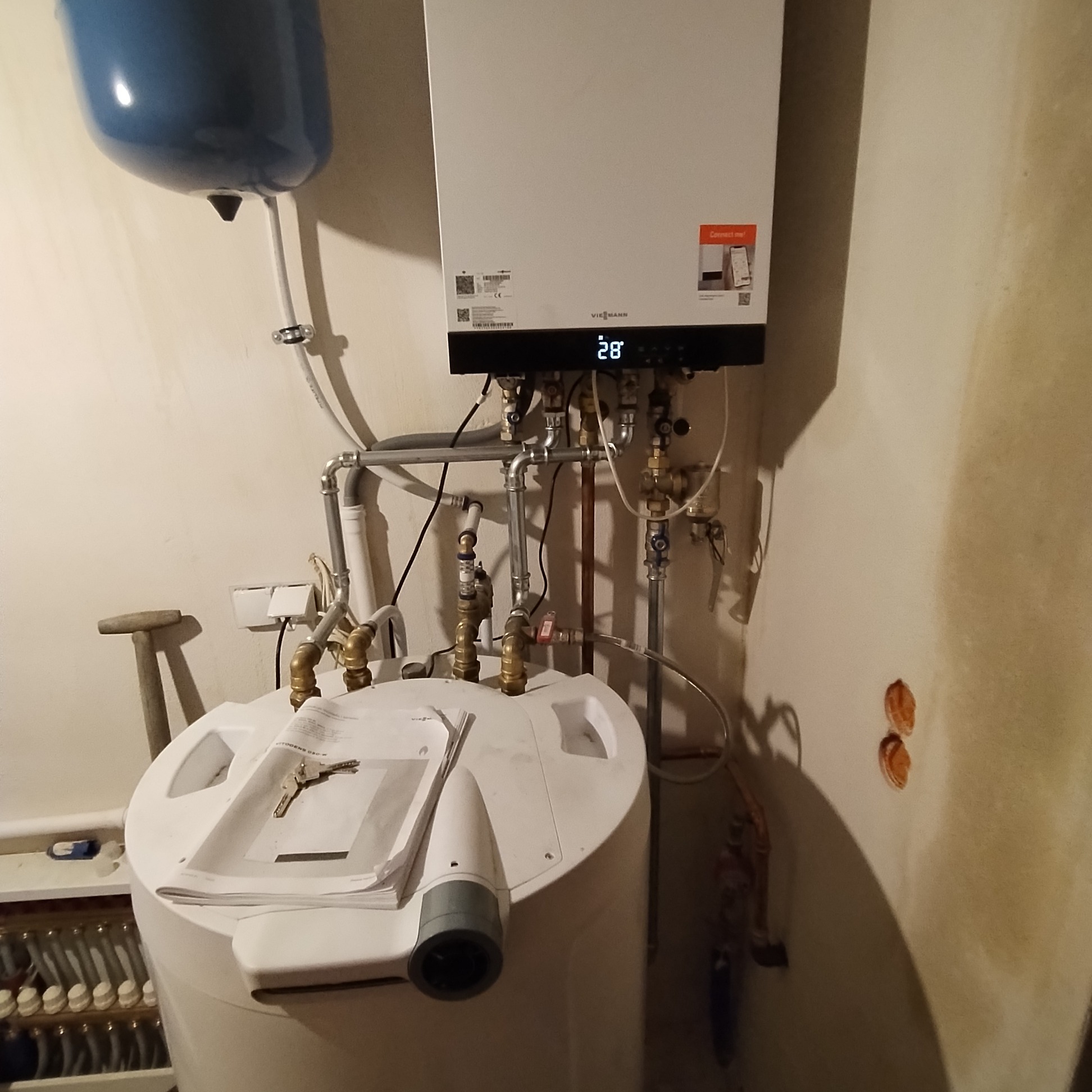 Nowoczesny piec gazowy Viessmann z podłączonym zasobnikiem ciepłej wody, widoczne rury i zawory, na zasobniku leżą dokumenty i klucze.