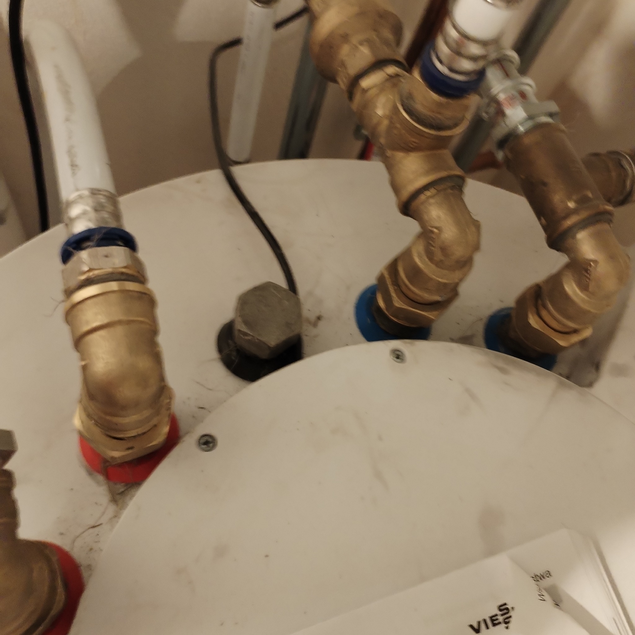 Szczegółowe ujęcie instalacji gazowej z mosiężnymi złączkami, białymi rurami i elementami z niebieskimi uszczelkami, zamontowanymi na białym zbiorniku, widoczne śruby i minimalne zabrudzenia.