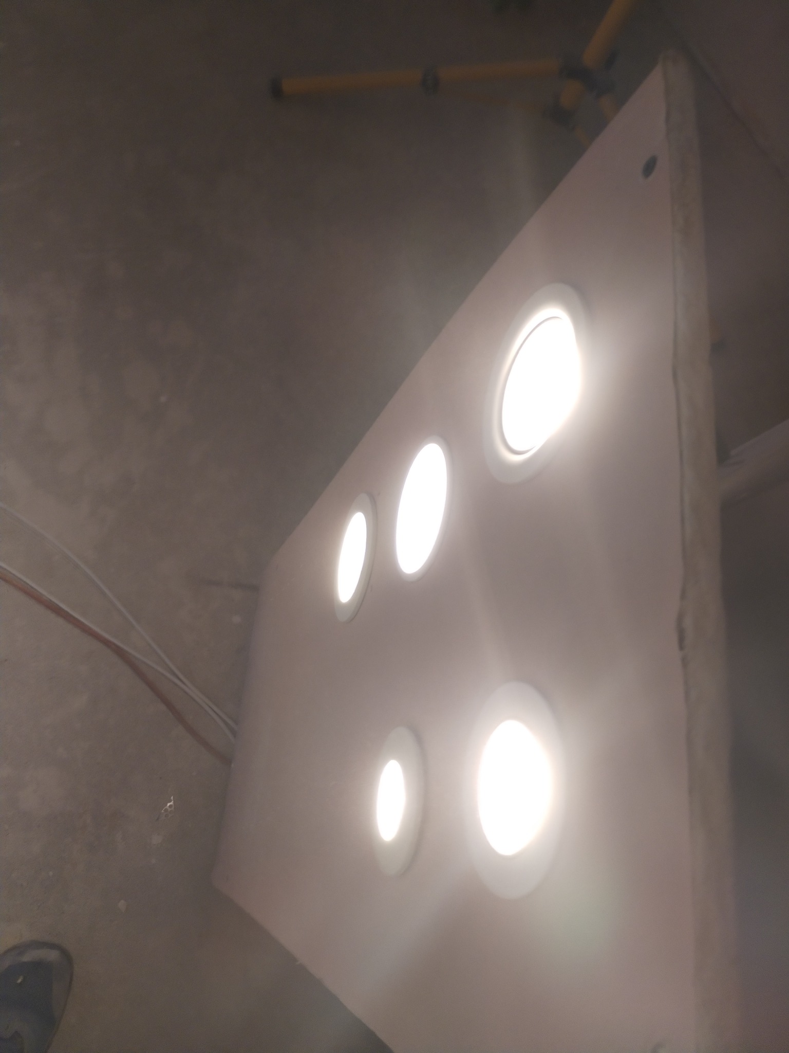 Panel z czterema okrągłymi otworami, z których wydobywa się intensywne światło, zamontowany na jasnej ścianie w surowym wnętrzu, widoczne przewody i fragment nogi statywu.