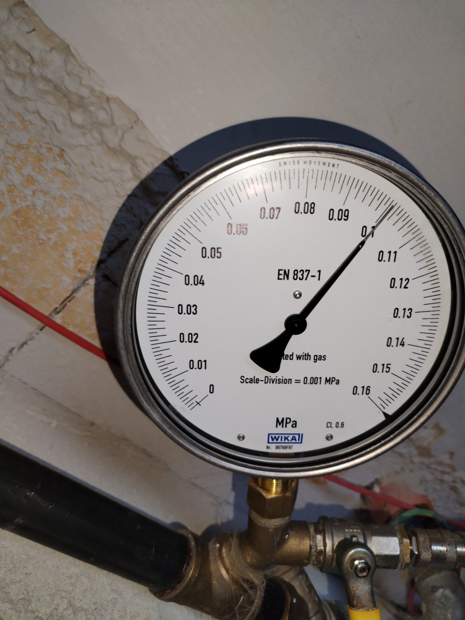 Manometr WIKA wskazujący ciśnienie gazu w instalacji, widoczne przyłącza rur i fragment ściany z nierówną fakturą.