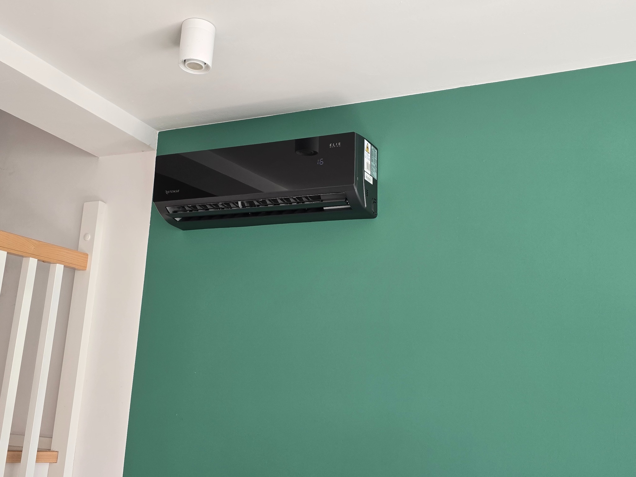 Czarny klimatyzator zamontowany na zielonej ścianie w pomieszczeniu z białym sufitem i elementami drewnianymi.