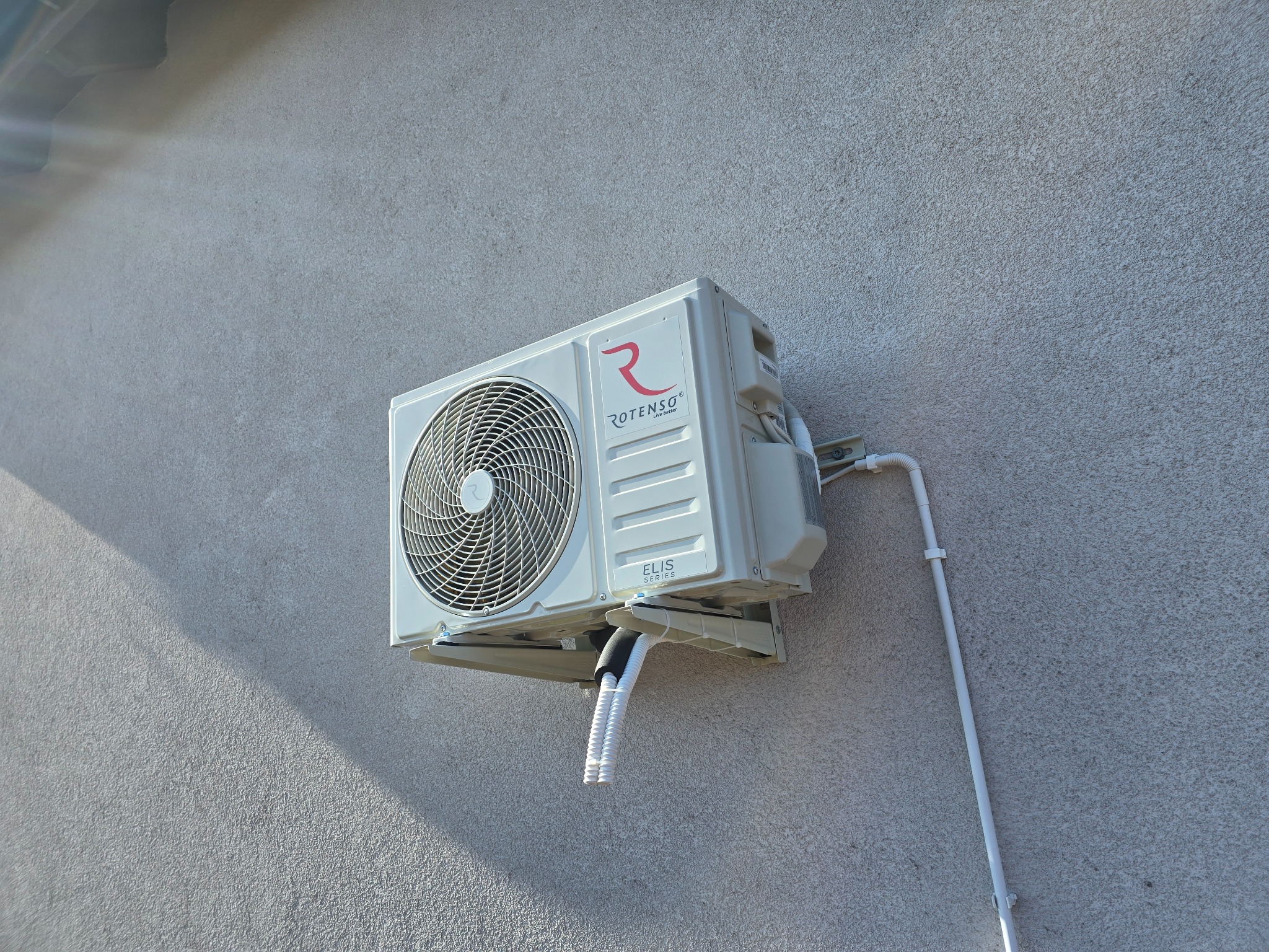 Zewnętrzna jednostka klimatyzacji Rotenso Elis Series zamontowana na szarej, teksturowanej ścianie budynku, widoczne białe rurki odprowadzające skropliny.