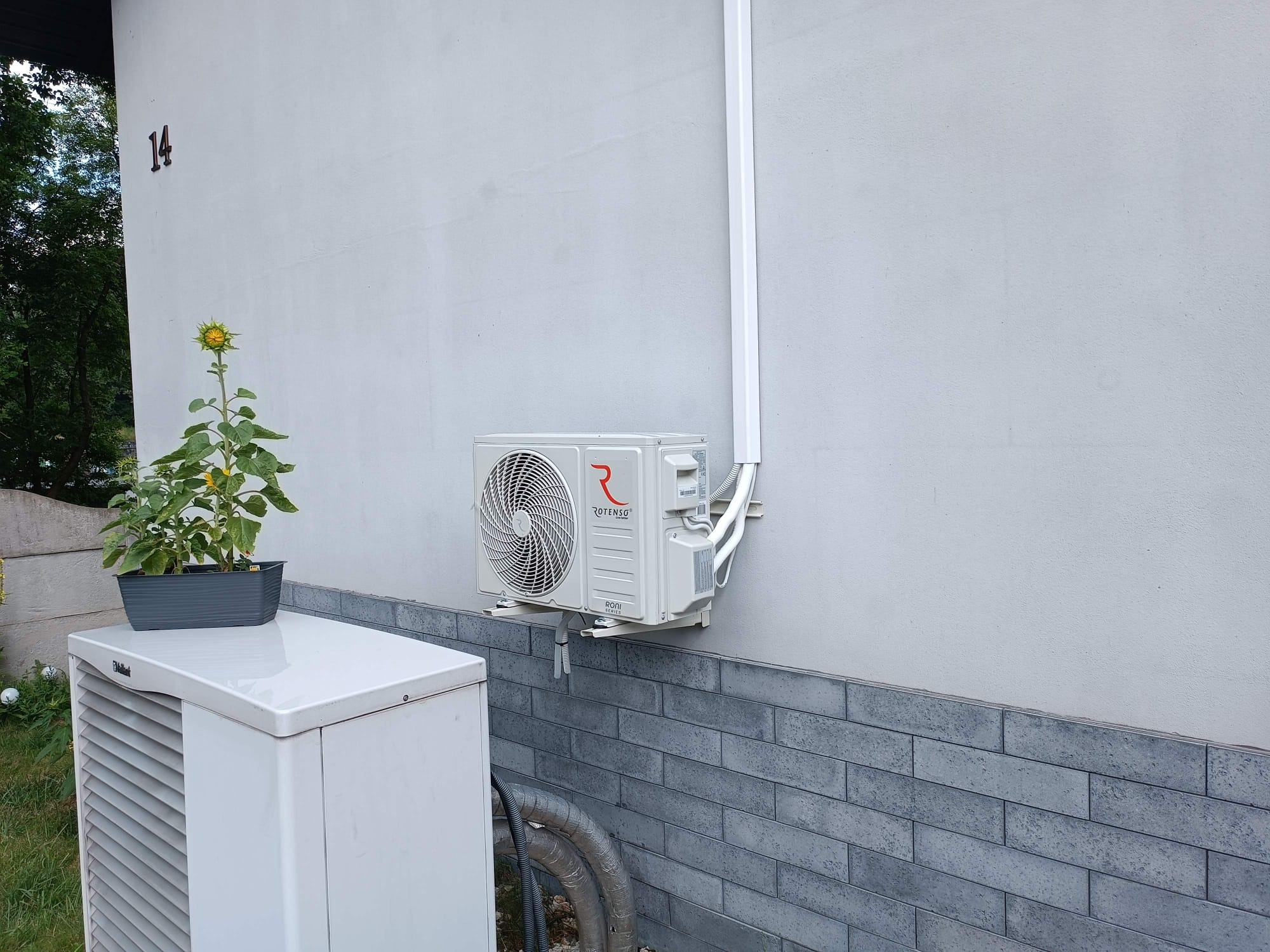 Zewnętrzna jednostka klimatyzacji Rotenso zamontowana na elewacji domu z dekoracyjną cegłą, obok jednostka zewnętrzna pompy ciepła Vaillant z doniczką ze słonecznikiem na górze.