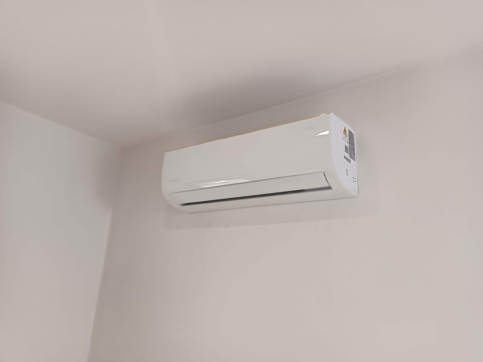 Biały klimatyzator ścienny z delikatnym złotym akcentem na górnej krawędzi, zamontowany na jasnej, gładkiej ścianie w rogu pomieszczenia.