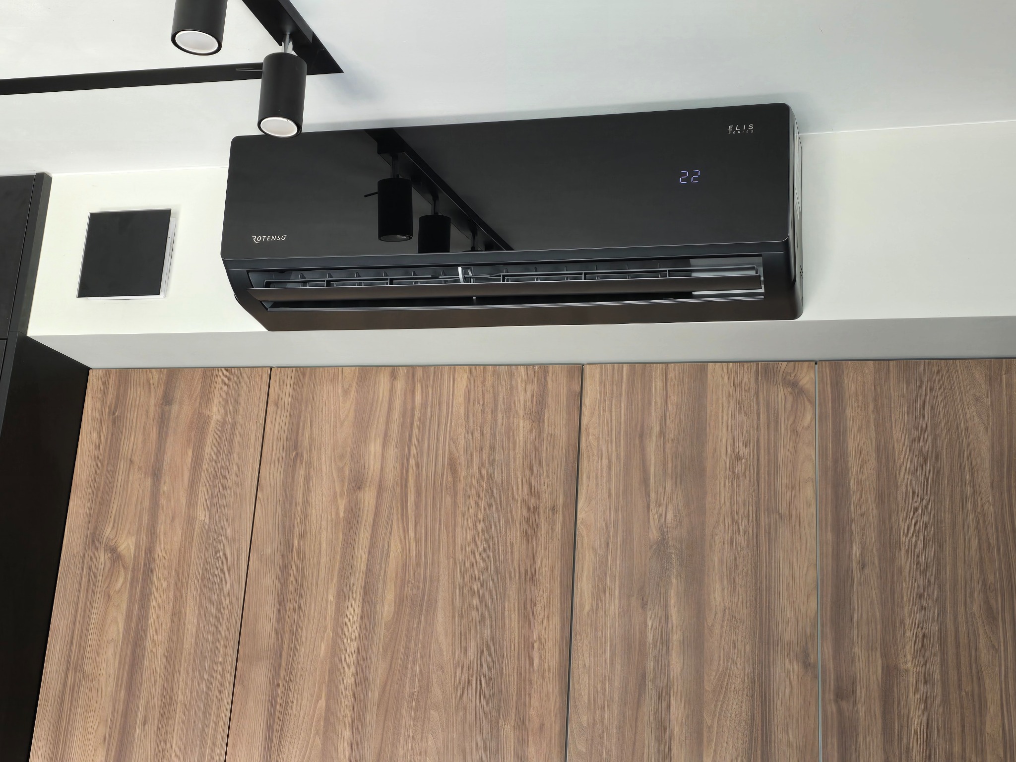Czarny klimatyzator Rotenso Elis Series zamontowany na białej ścianie nad drewnianymi szafkami, wyświetlający temperaturę 22 stopnie.