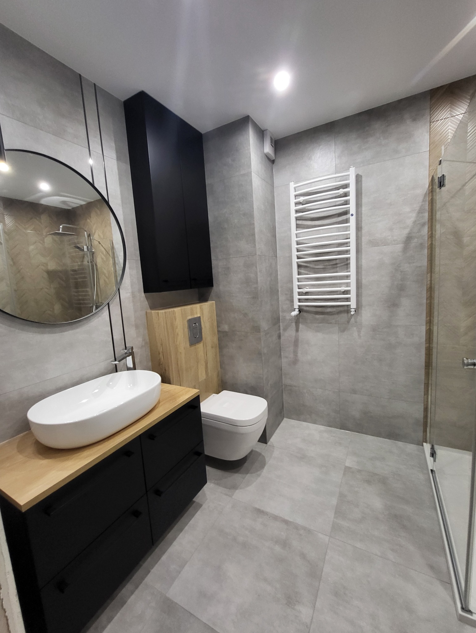 Nowoczesna łazienka z szarymi płytkami, wiszącą szafką z umywalką na drewnianym blacie, toaletą podwieszaną i grzejnikiem drabinkowym.