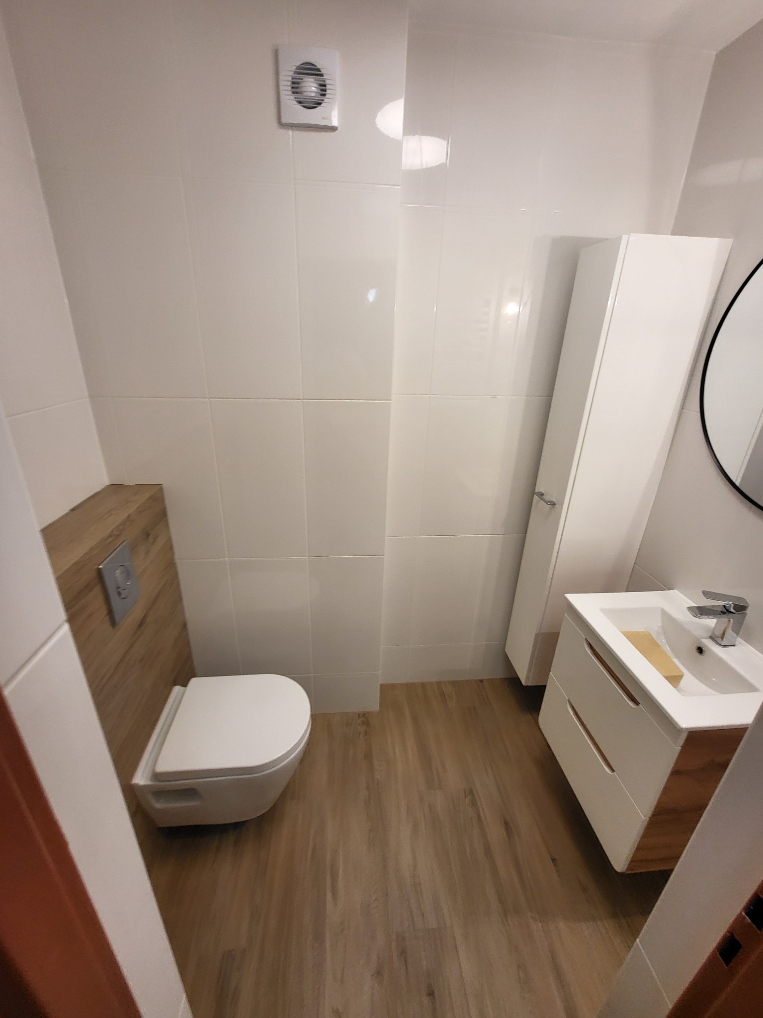 Nowoczesna łazienka z białymi płytkami, wiszącą toaletą, szafką pod umywalkę z drewnianymi akcentami i drewnopodobną podłogą, widoczny wentylator sufitowy i okrągłe lustro.