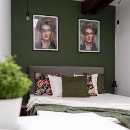 Bartlomiej Skorupski - Sypialnia z łóżkiem z szarym zagłówkiem, poduszkami w kwiaty i zielenią, ciemnozielona ściana z dwoma portretami w czarnych ramkach i wiszącymi żarówkami.