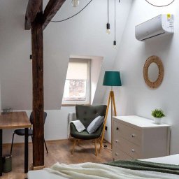 Bartlomiej Skorupski - Jasna sypialnia na poddaszu z drewnianą belką konstrukcyjną, lampami na kablach zwisających z sufitu, klimatyzatorem, komodą, fotelem i biurkiem