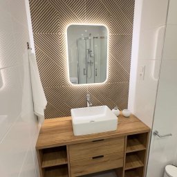 Bartlomiej Skorupski - Nowoczesna łazienka z drewnianą szafką pod umywalką, białą umywalką w kształcie prostokąta, podświetlanym lustrem i ścianą z geometrycznym wzorem, widoczna kabina prysznicowa w odbiciu lustra.