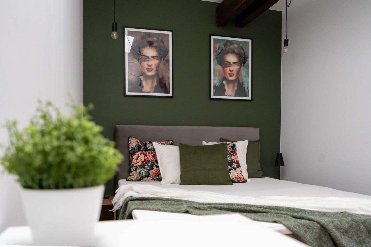 Sypialnia z łóżkiem z szarym zagłówkiem, poduszkami w kwiaty i zielenią, ciemnozielona ściana z dwoma portretami w czarnych ramkach i wiszącymi żarówkami.