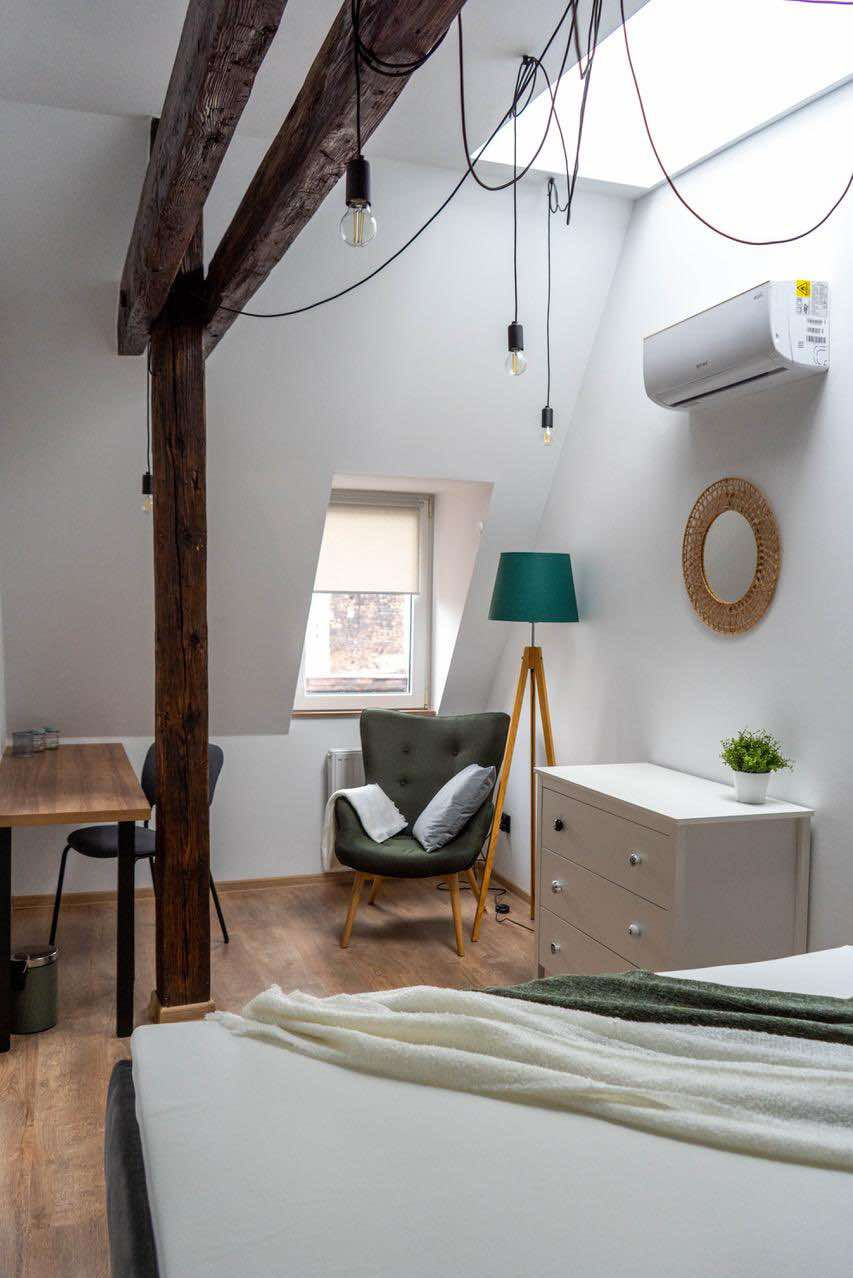 Jasna sypialnia na poddaszu z drewnianą belką konstrukcyjną, lampami na kablach zwisających z sufitu, klimatyzatorem, komodą, fotelem i biurkiem