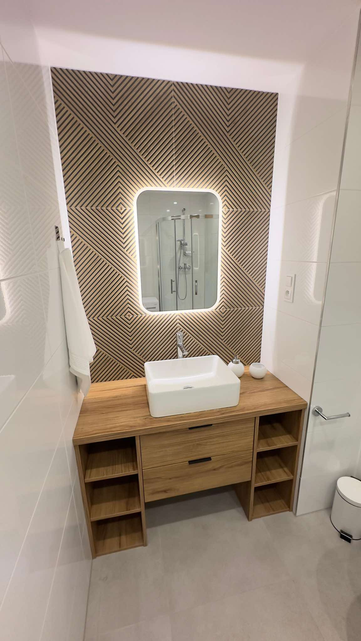 Nowoczesna łazienka z drewnianą szafką pod umywalką, białą umywalką w kształcie prostokąta, podświetlanym lustrem i ścianą z geometrycznym wzorem, widoczna kabina prysznicowa w odbiciu lustra.