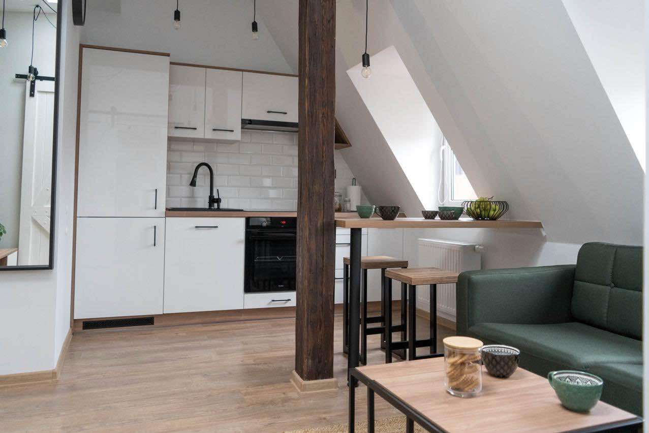 Jasna kuchnia z białymi szafkami i czarnymi akcentami, drewniana belka konstrukcyjna, stół barowy z hokerami oraz fragment salonu z sofą w tle, poddasze.