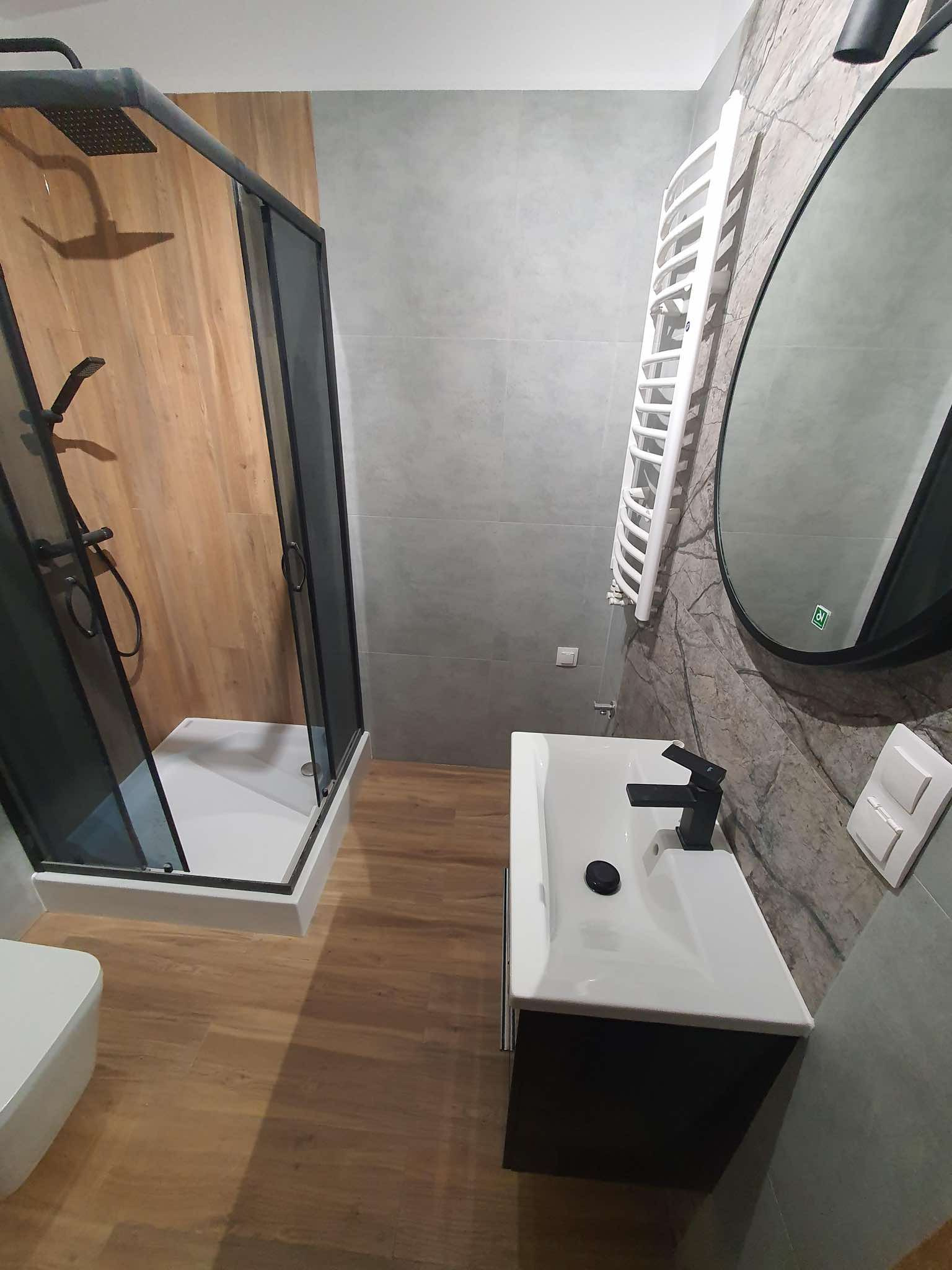 Nowoczesna łazienka w Chorzowie z kabiną prysznicową typu walk-in z czarnymi ramami, białą umywalką z czarną armaturą, grzejnikiem drabinkowym i okrągłym lustrem.