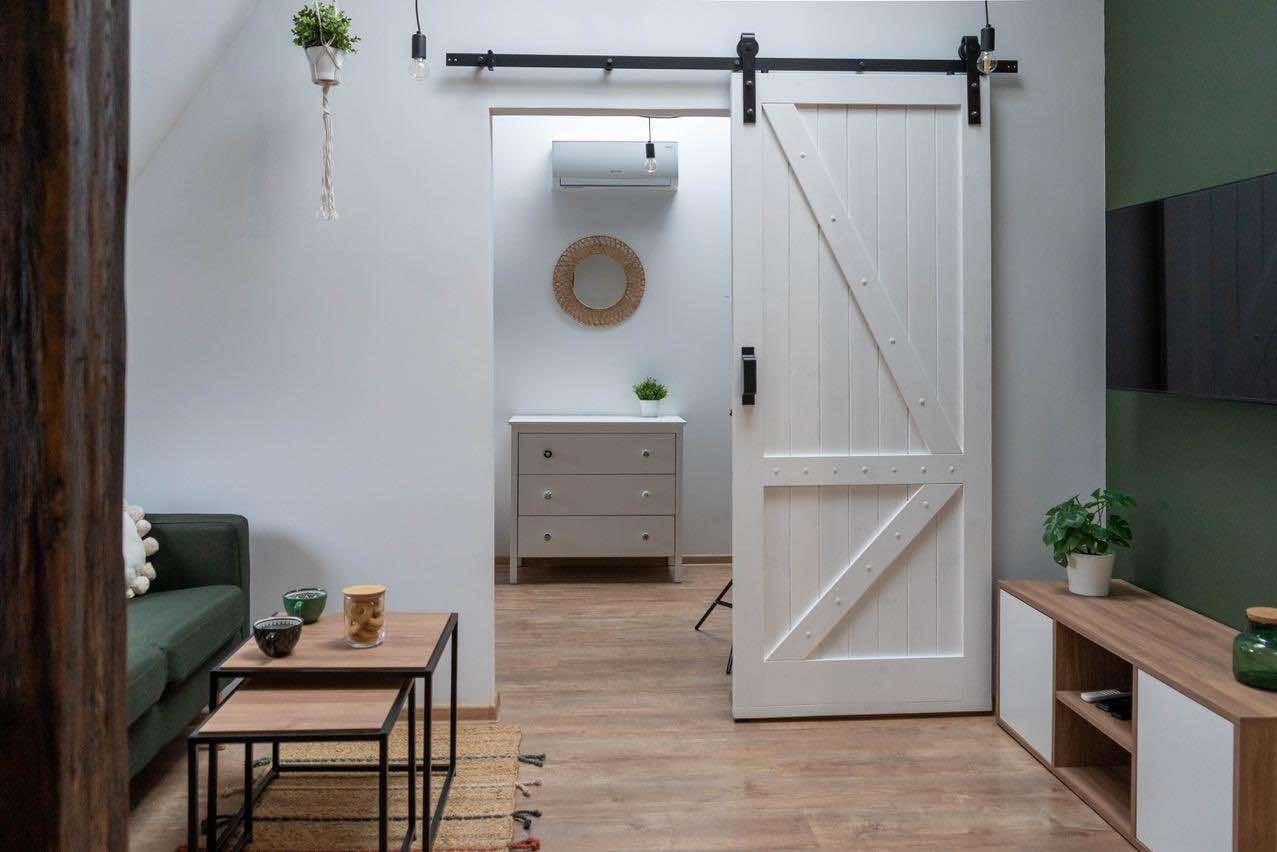 Białe, przesuwne drzwi stodołowe w stylu rustykalnym z czarnym okuciem, częściowo otwarte, widok na wnętrze z komodą, klimatyzatorem i okrągłym lustrem, fragment salonu z zieloną sofą i stolikiem...