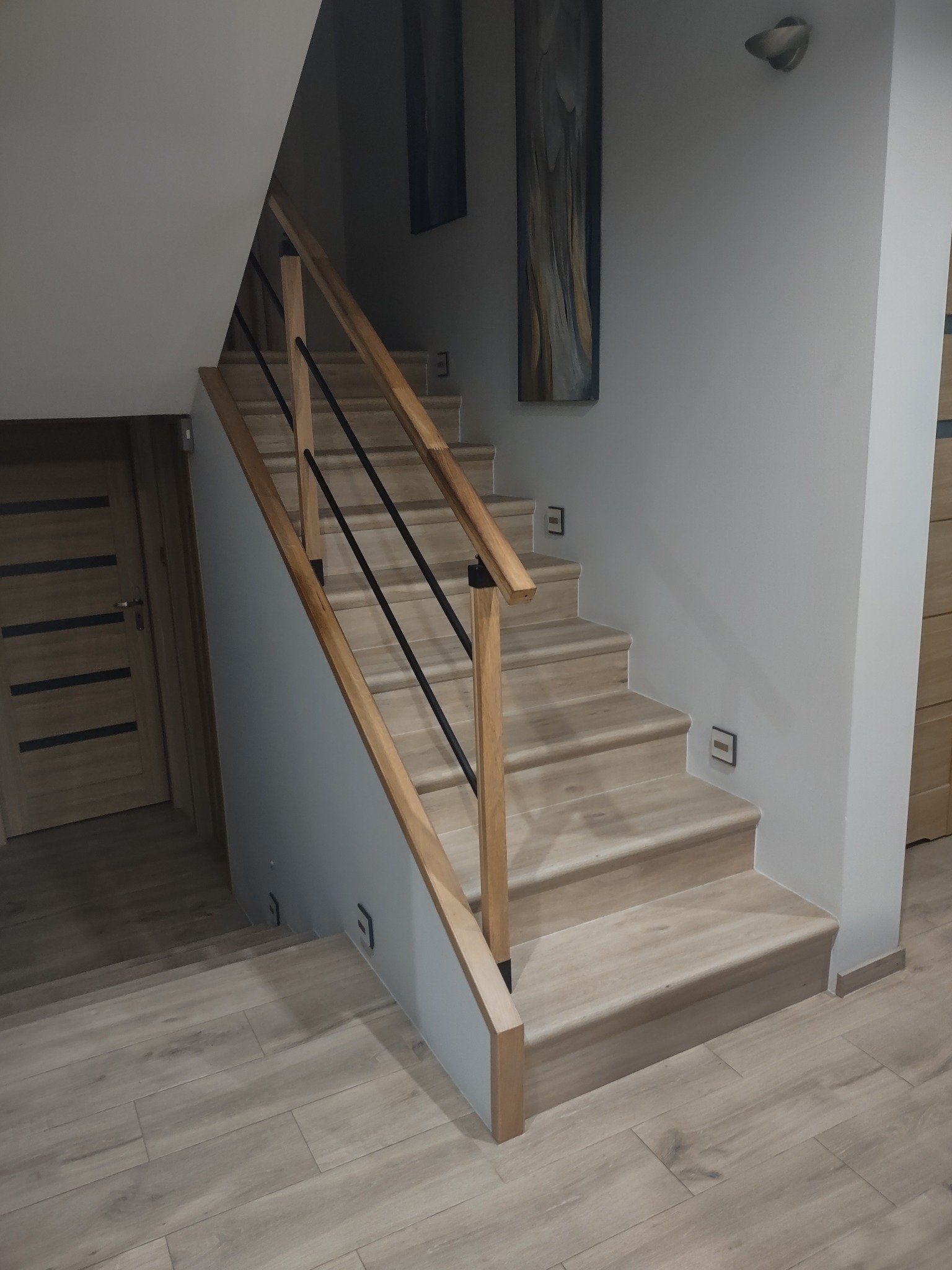 Schody z drewnianymi stopniami i czarnymi metalowymi prętami balustrady, oświetlenie schodowe wbudowane w ścianę, podłoga wyłożona panelami imitującymi drewno.