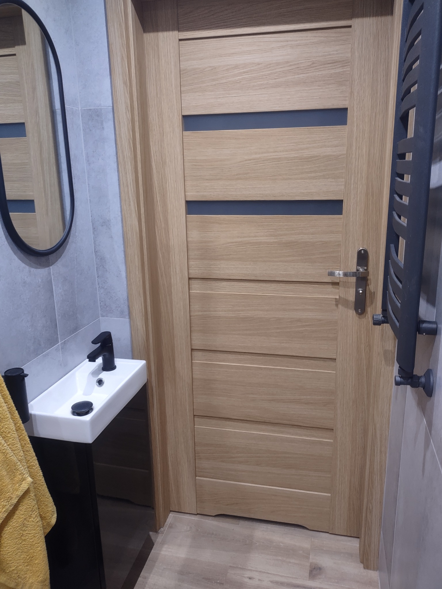 Nowoczesna łazienka z umywalką na czarnym postumencie, owalnym lustrem w czarnej ramie, drewnianymi drzwiami z ciemnymi wstawkami i szarym grzejnikiem drabinkowym.