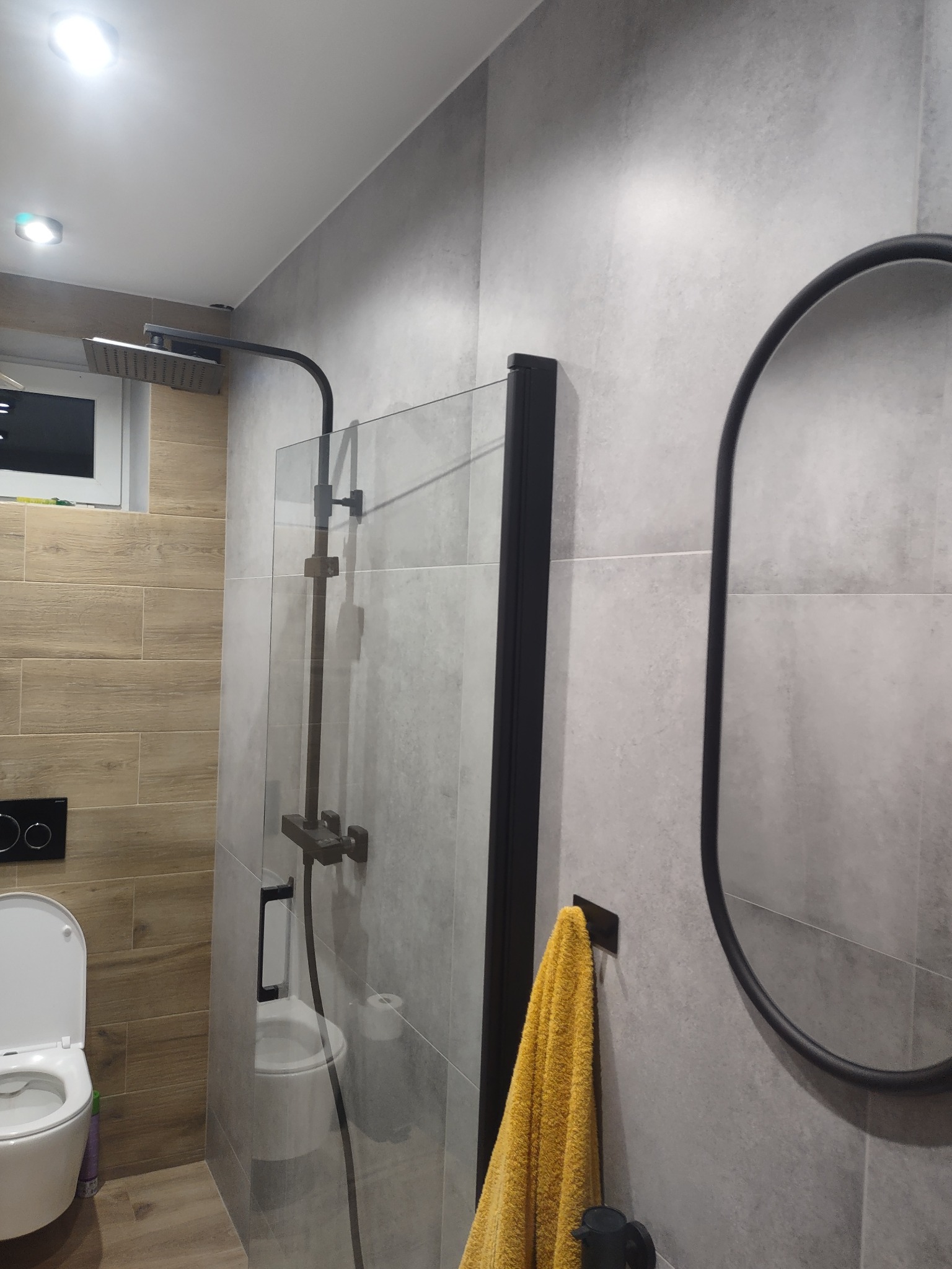 Nowoczesna łazienka z szarymi płytkami, kabiną prysznicową typu walk-in z czarnymi elementami, owalnym lustrem w czarnej ramie i żółtym ręcznikiem.