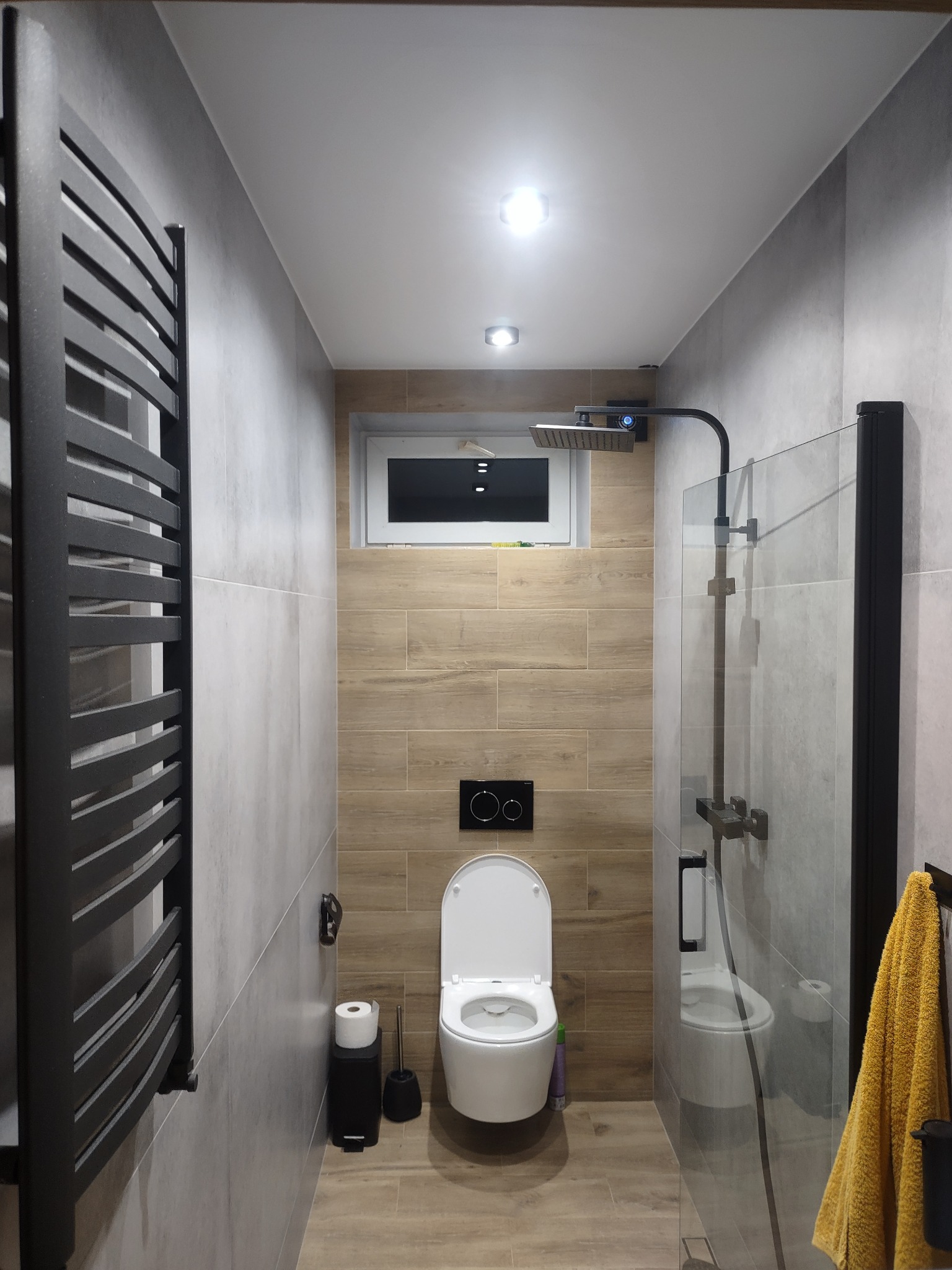 Nowoczesna łazienka z szarymi i drewnopodobnymi płytkami, czarną armaturą, grzejnikiem drabinkowym i kabiną prysznicową typu walk-in.