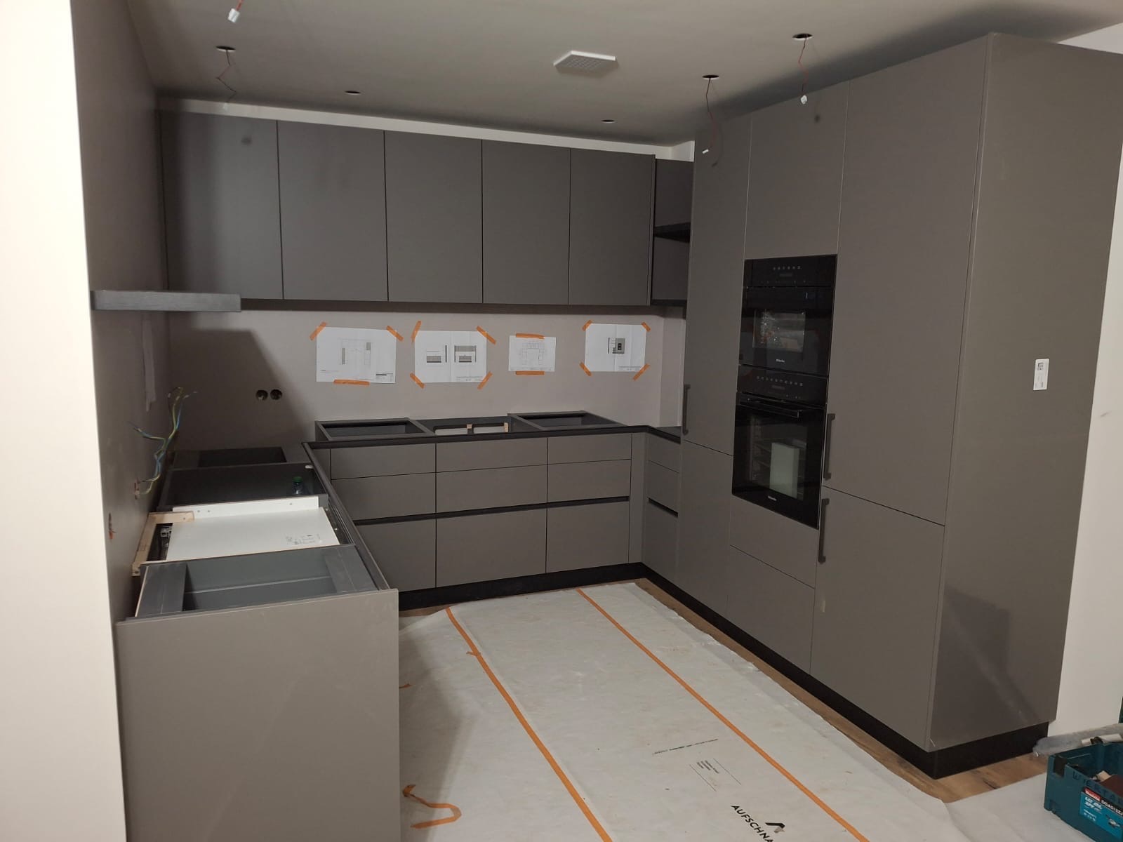 Szara kuchnia w trakcie montażu, widoczne szafki bez frontów, piekarnik Miele, plany na ścianie, zabezpieczona podłoga i wystające kable.