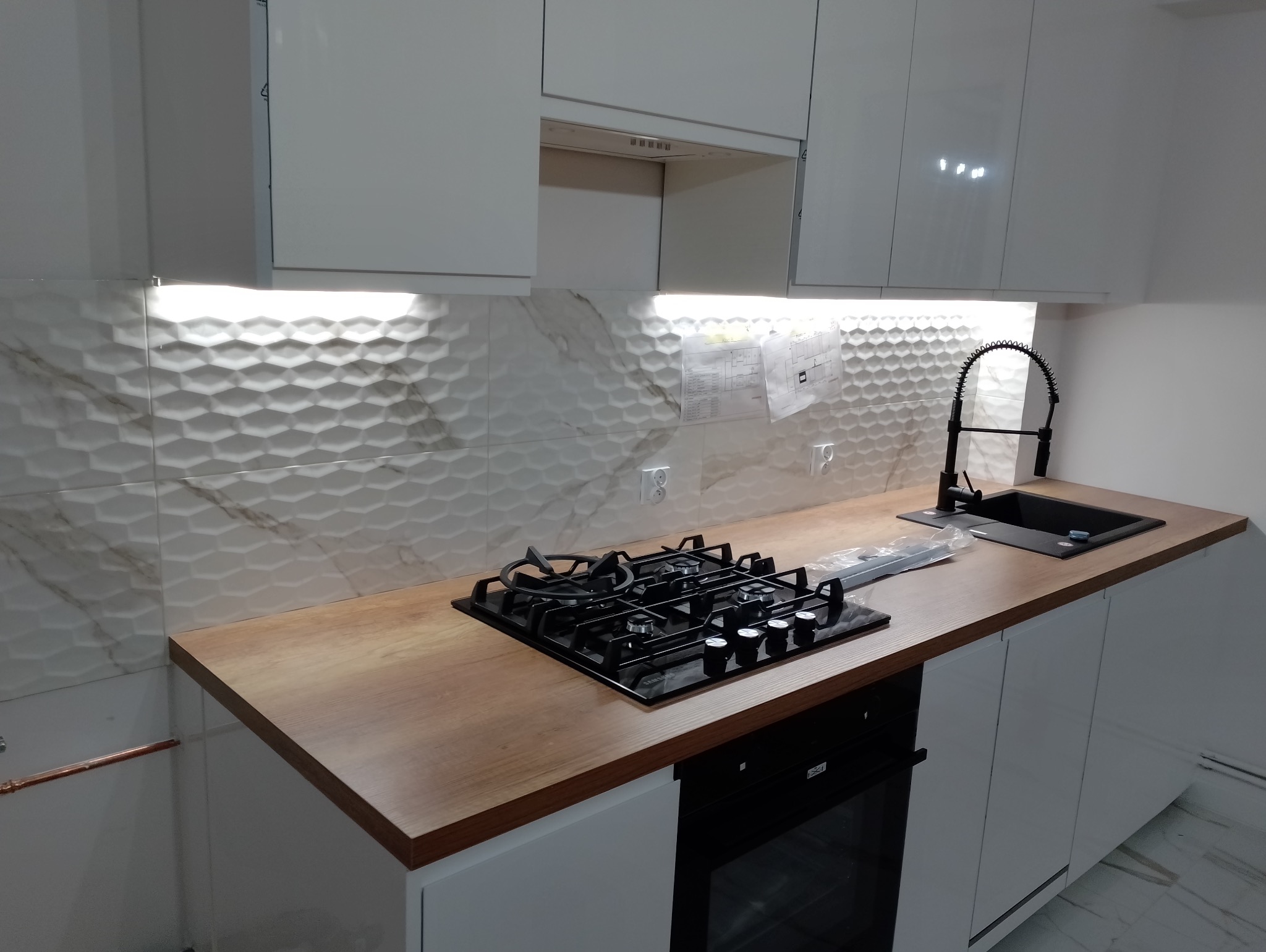 Nowoczesna, biała kuchnia z drewnianym blatem, płytą gazową i czarnym zlewem z baterią na sprężynie, wykończona płytkami 3D imitującymi marmur, oświetlenie podszafkowe, widoczne plany instalacji...