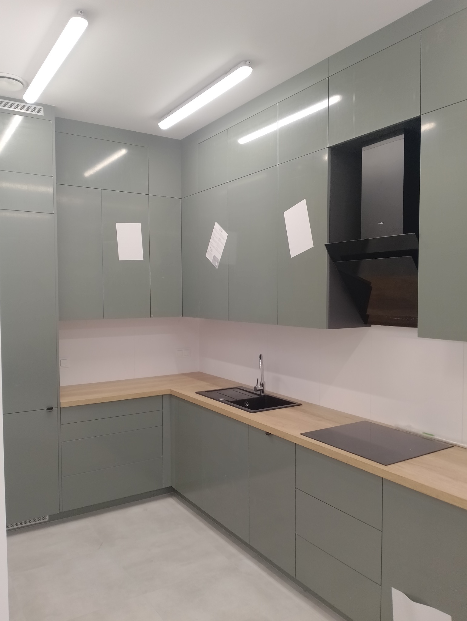 Nowoczesna kuchnia z szarymi, lakierowanymi szafkami, drewnianym blatem i czarnym okapem Amica, z widocznymi kartkami na frontach szafek.