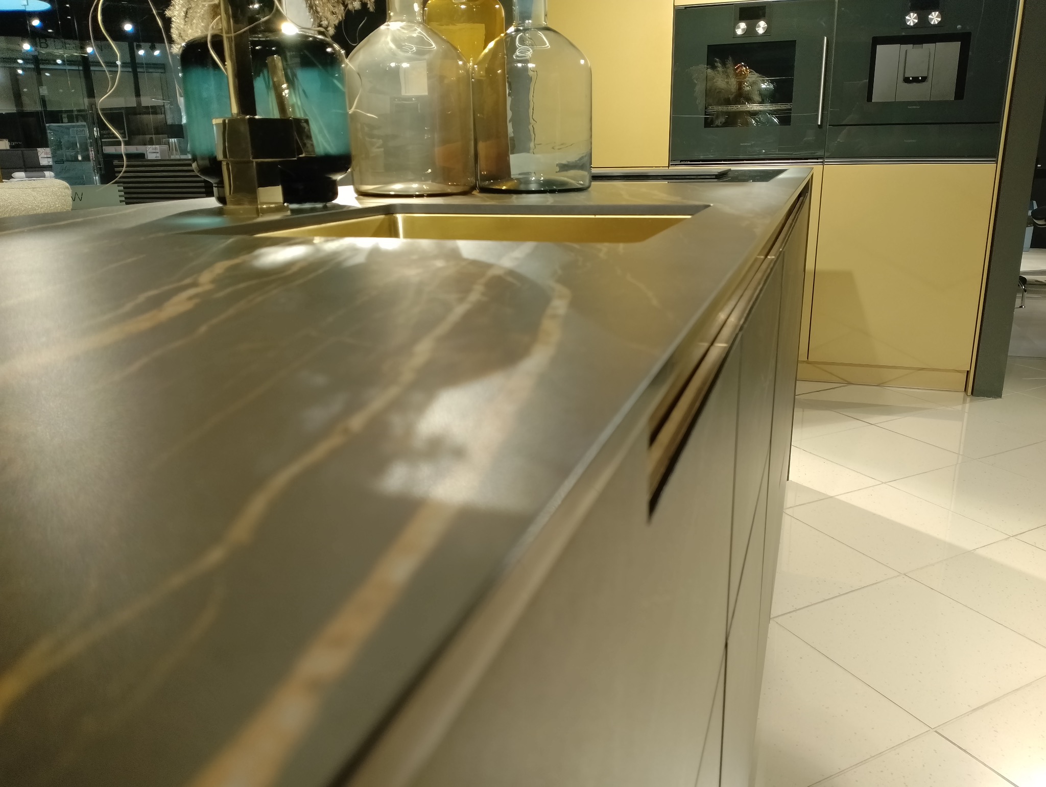 Błyszczący, ciemny blat kuchenny z marmurkowym wzorem, zintegrowany z metalowym zlewem, na którym stoją dekoracyjne szklane butelki. W tle zabudowa kuchenna z piekarnikiem i szafkami w odcieniach...