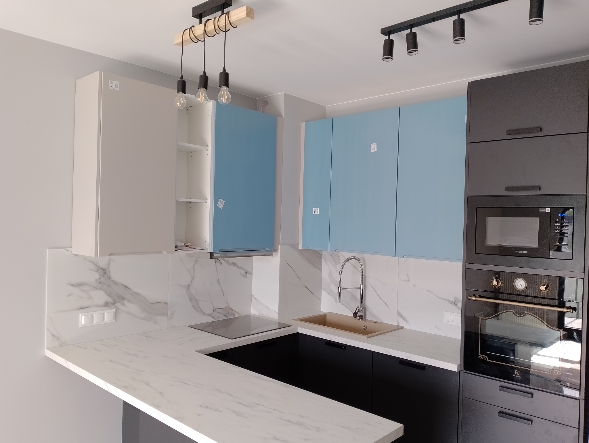 Nowoczesna kuchnia z białym blatem imitującym marmur, jasnoniebieskimi i grafitowymi szafkami, oraz oświetleniem punktowym i wiszącymi lampami z dekoracyjnymi żarówkami.