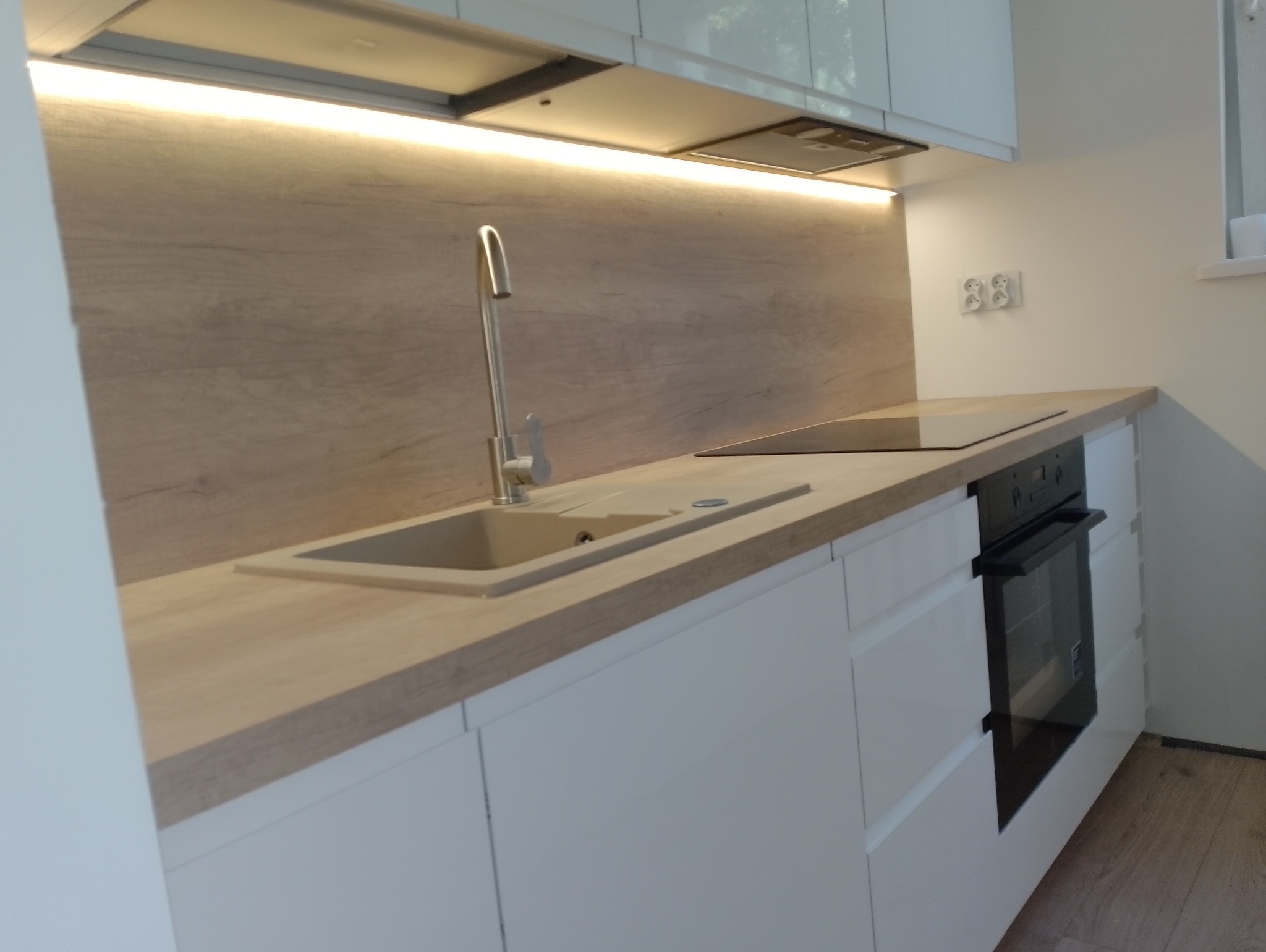 Nowoczesna, jasna kuchnia z drewnianym blatem, białymi szafkami i beżowym zlewem podświetlona listwą LED. Widoczny piekarnik elektryczny i płyta indukcyjna.