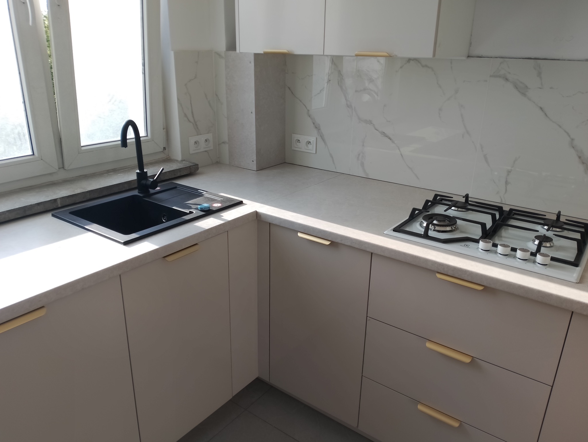 Nowoczesna, jasna kuchnia z beżowymi szafkami, złotymi uchwytami, czarnym zlewem pod oknem i białą płytą gazową na tle marmurowych płytek ściennych.