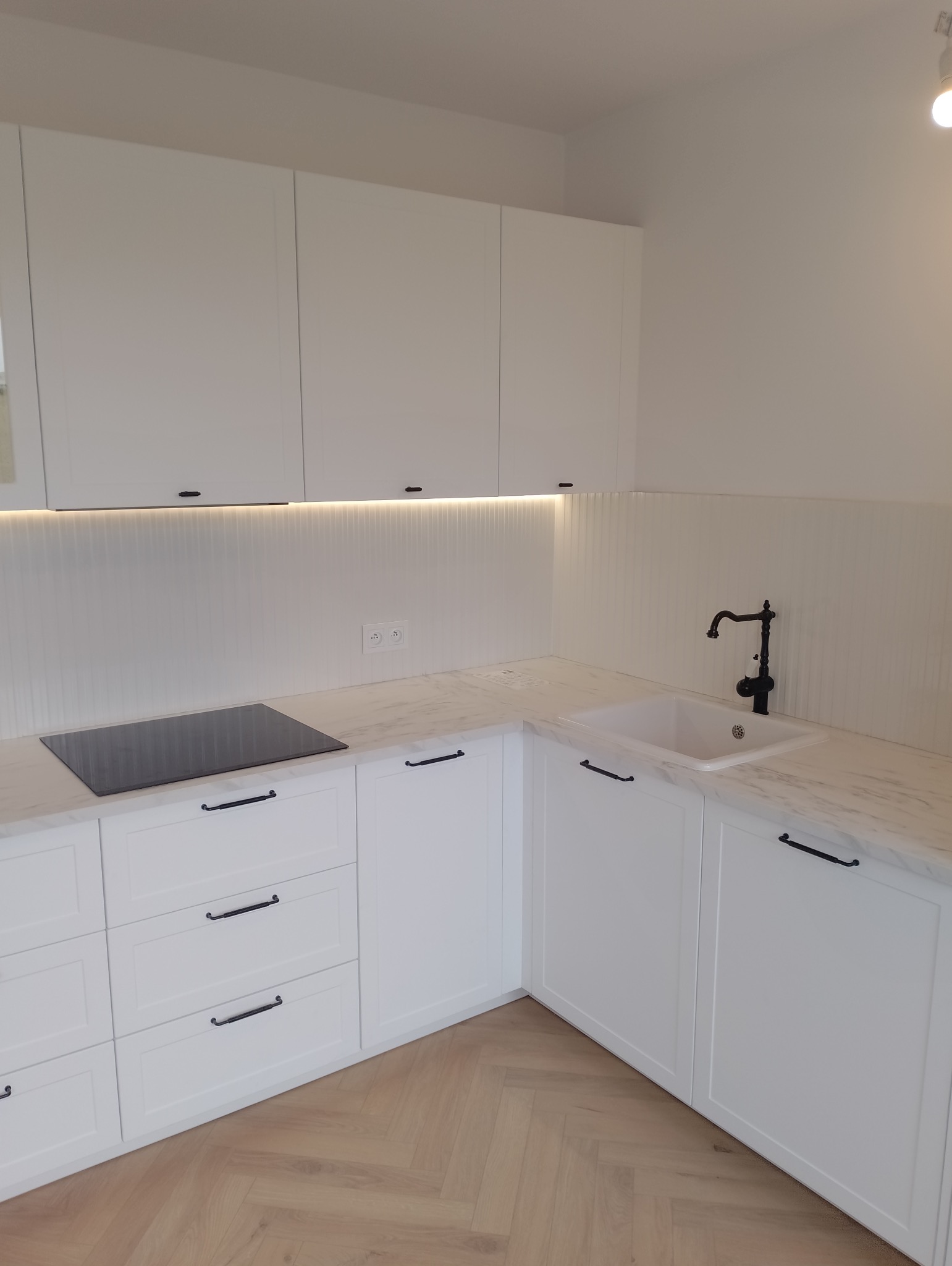 Nowoczesna biała kuchnia z marmurowym blatem, czarną armaturą i oświetleniem LED pod szafkami górnymi, parkiet w jodełkę.