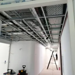 Korytarz w trakcie prac wykończeniowych, widoczne metalowe profile sufitowe z instalacjami, drabina rozstawiona w głębi, narzędzia i skrzynki na pierwszym planie, ściany przygotowane do malowania.