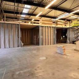 Jakub Zborowski - Wnętrze hali w trakcie budowy z metalowymi konstrukcjami ścian działowych wypełnionymi materiałem izolacyjnym, widoczna podnośnik nożycowy z pracownikiem na górze, surowe betonowe podłoże...